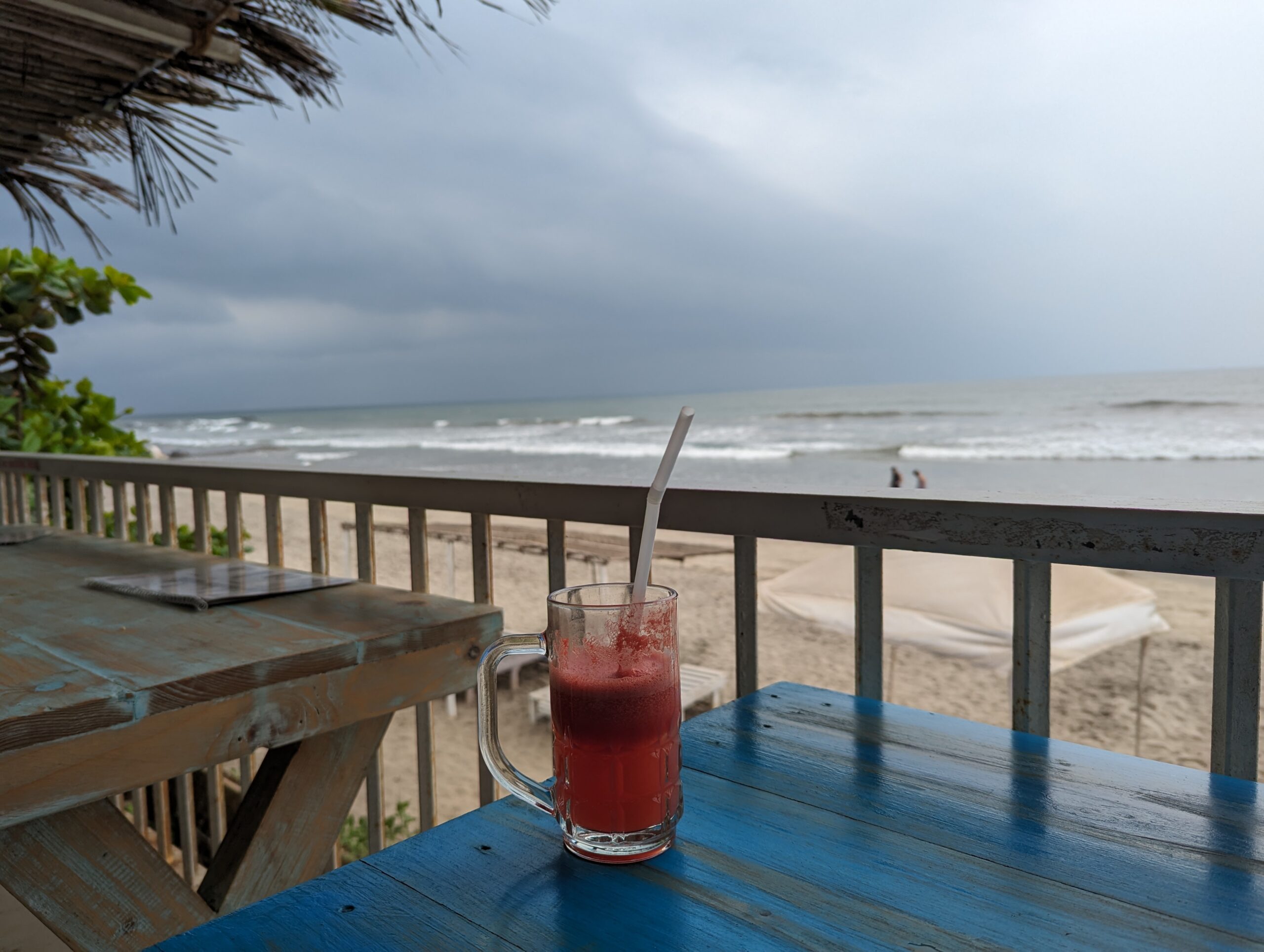 A picture of a glass of watermelon juice in the foreground with a beach in Goa in the backdrop. There are stormy clouds in the background.