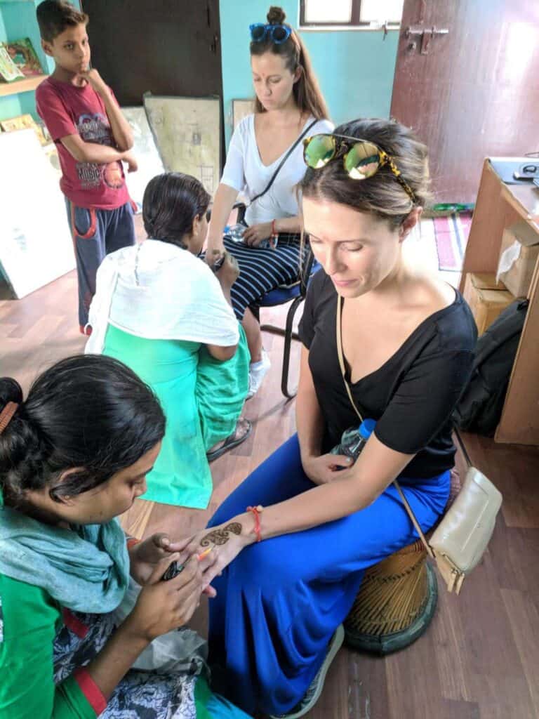 A woman sitting in a small room gets a traditional henna design applied on her hand by a local artist. The artist carefully works on the intricate pattern, while another woman beside her also receives henna. A young boy and other individuals observe the process in the background. The setting is simple, with a relaxed and cultural atmosphere, showcasing a traditional art form in India.