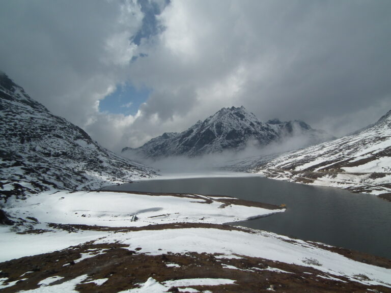 A high Himalayan lake in the mountains which is surrounded by snow clad mountains. It is half frozen with big clouds in the backdrop.