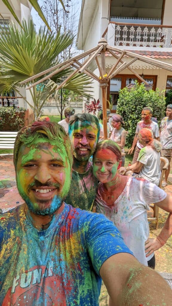 A selfie of 3 people with colorful green, red, blue and pink powder all over their faces celebrating Holi. There are other people in the background covered in similar powder colors. 