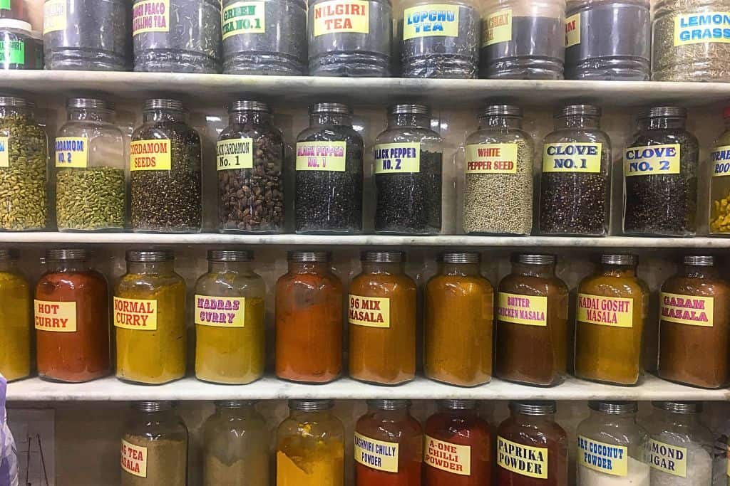 A display of various spices and teas in glass jars, neatly arranged on shelves in an Indian spice shop. Labels on the jars include a variety of spices like cardamom, black pepper, white pepper, clove, curry powders, garam masala, and masala mixes. Some tea varieties like green tea, Lopchu tea, and Nilgiri tea are also visible, offering a vibrant assortment of flavors and aromas.