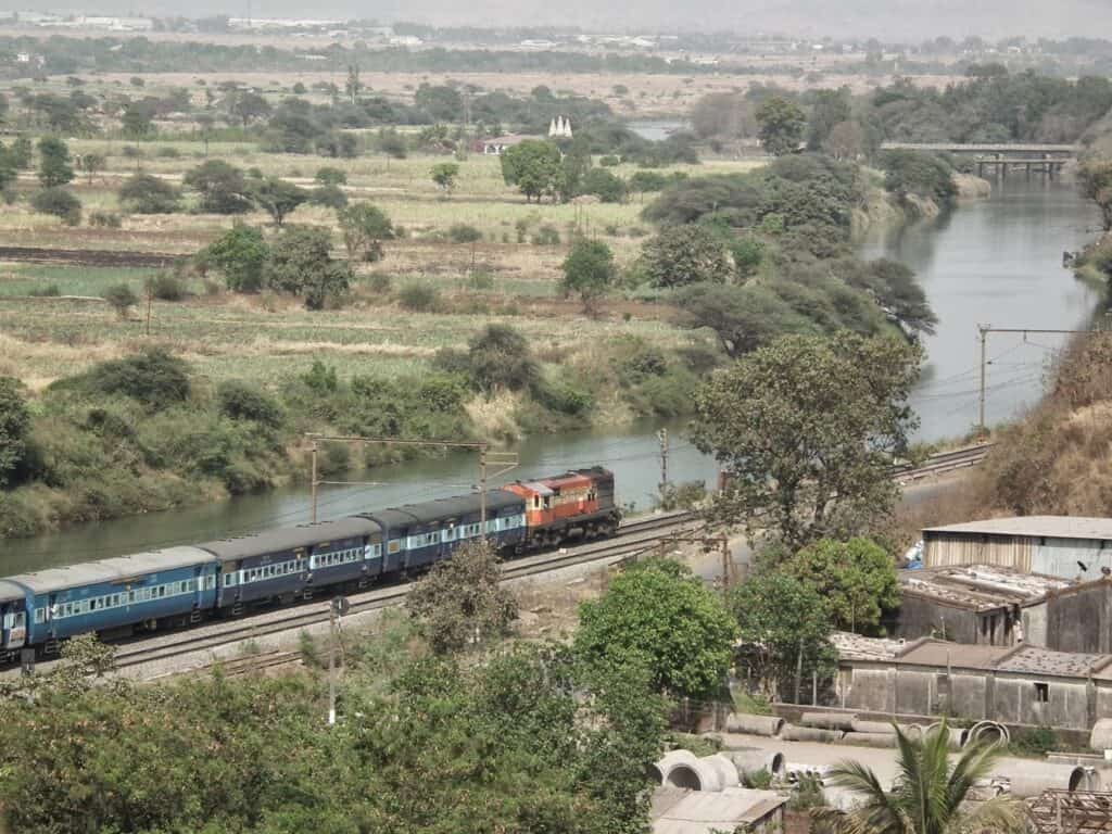 An aerial view of a rural Indian landscape with a train passing along tracks beside a winding river. The scene includes green fields, scattered trees, and small buildings in the distance. The train has blue carriages and an orange locomotive.