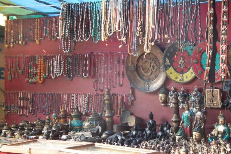 A vibrant market stall displaying an array of traditional Indian handicrafts, including colorful beaded necklaces, brass trinkets, Buddhist statues, ornate teapots, and decorative wall plates with spiritual symbols like "Om" and Buddha motifs. The items are neatly arranged, creating an enticing and cultural showcase of local craftsmanship.