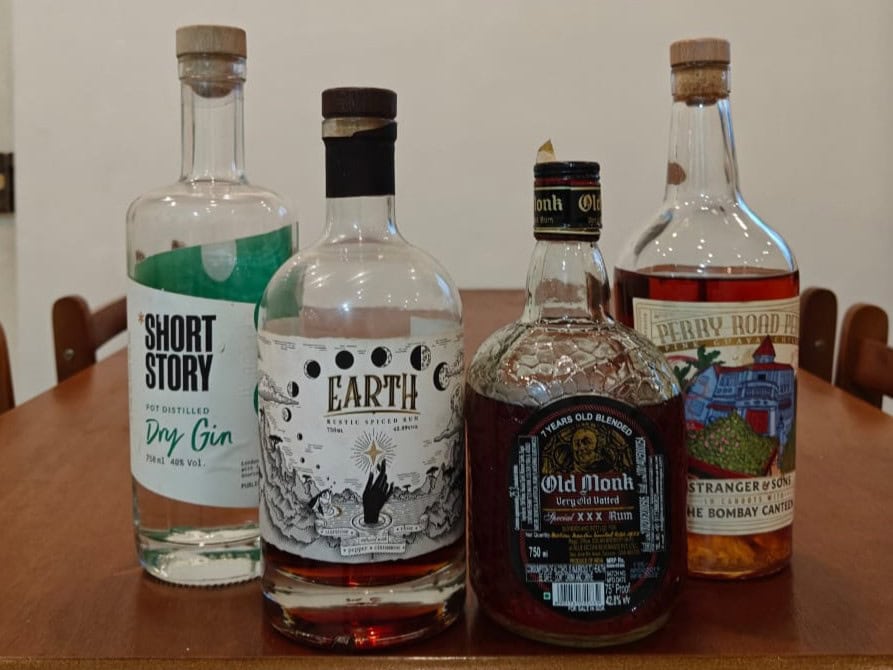 Four bottles of Indian spirits on a wooden table: Short Story Dry Gin, Earth Exotic Spiced Rum, Old Monk Rum, and Stranger & Sons Perry Road Peru