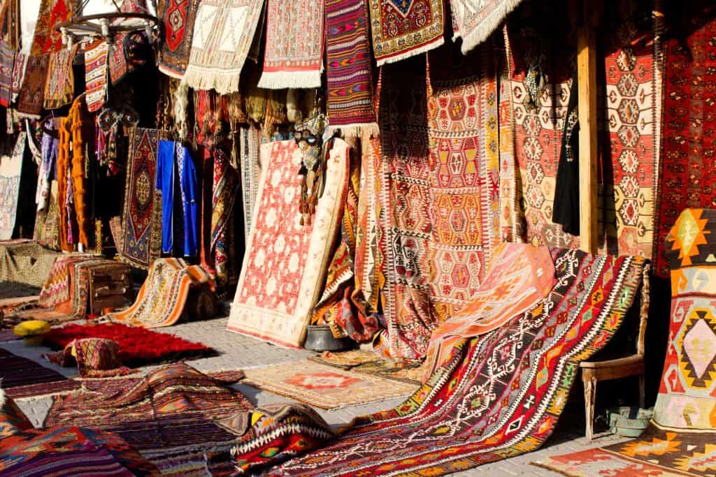 A colorful outdoor display of traditional Kashmiri rugs, showcasing intricate patterns and designs. The vibrant textiles are draped over stalls and spread across the ground, featuring a variety of rich reds, oranges, blues, and geometric motifs. Some of the rugs hang vertically, while others are laid out flat, creating a vibrant tapestry of handwoven artistry