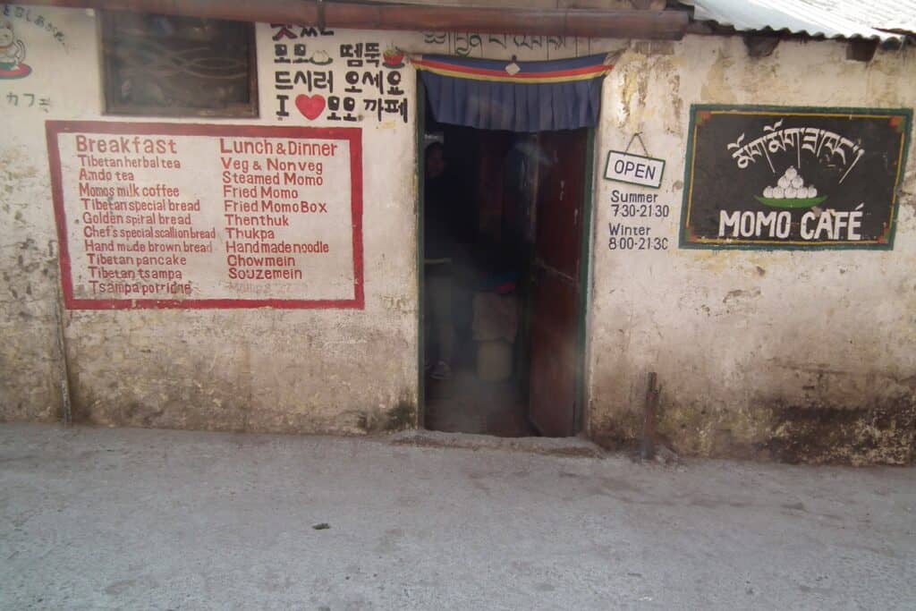 A picture of the outside of a small Tibetan cafe in Mcleodganj, India. There is a small door in the centre with a painted menu on the left with a sign that says momo cafe to the right of the door. 