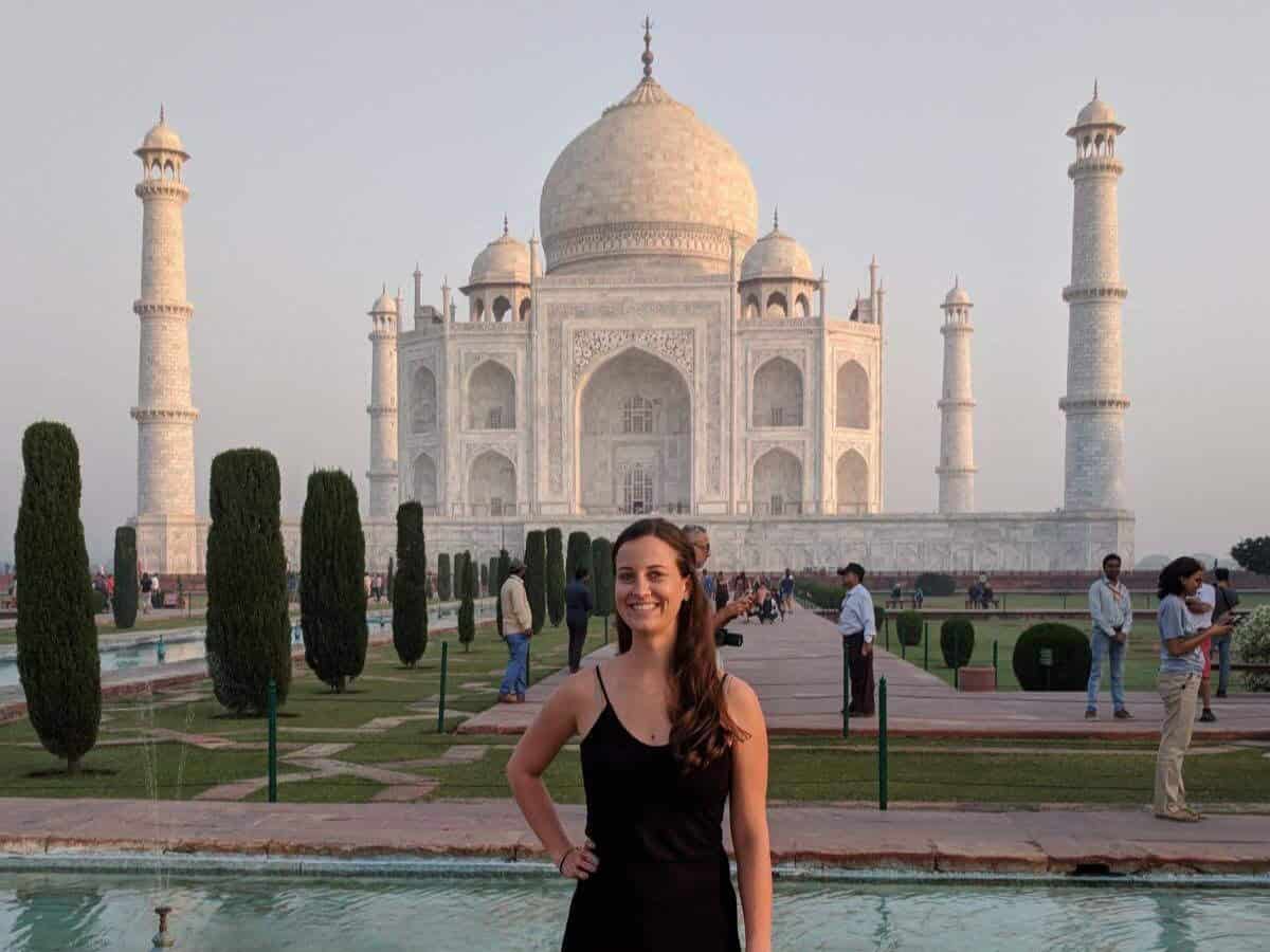 Girl in plain black dress posing by the reflecting pool in front of Taj Mahal, with other tourists in the background.