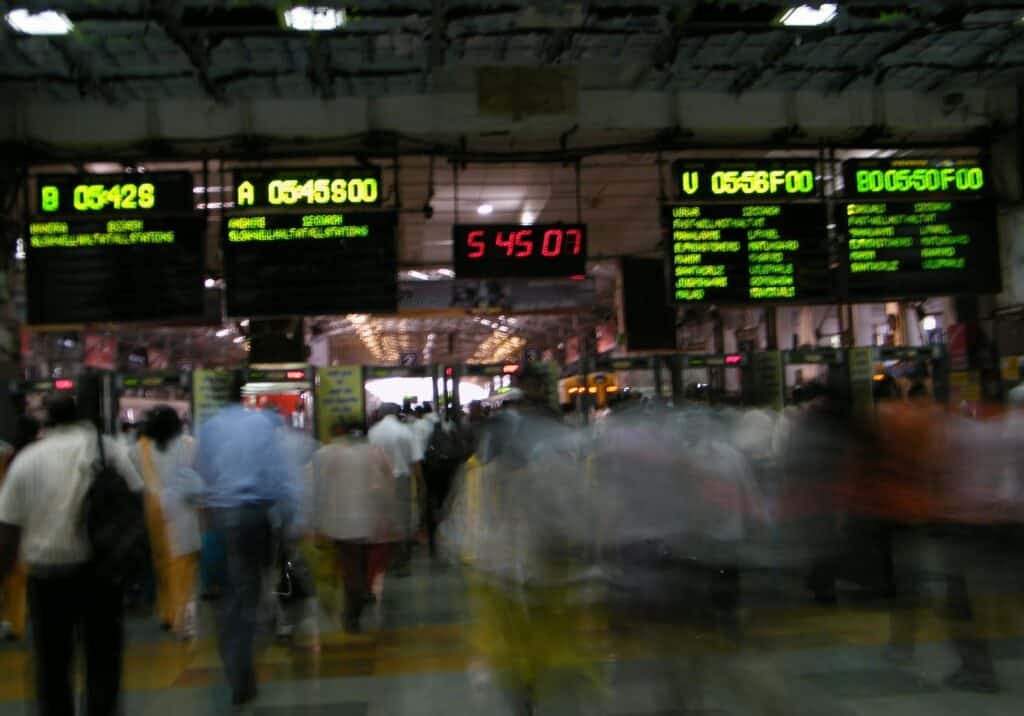 A crowd of people during the evening rush hour at the Churchgate train station in Mumbai. The photographer has used a slow shutter speed making the crowd blurred with signs displaying outbound trains in the background. There is a giant digital clock that hangs in he middle with the time reading 5.45.07