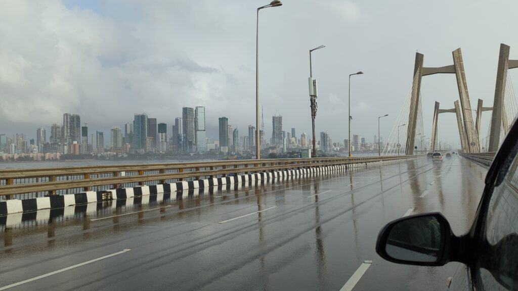 A picture of the Bandra-Worli sea link in Mumbai taken from inside a car. You can see the pillars of the bridge, a drenched road surface and the towers in the background with a glimpse of the sea. You can also see the side view mirror and the front left window of the car