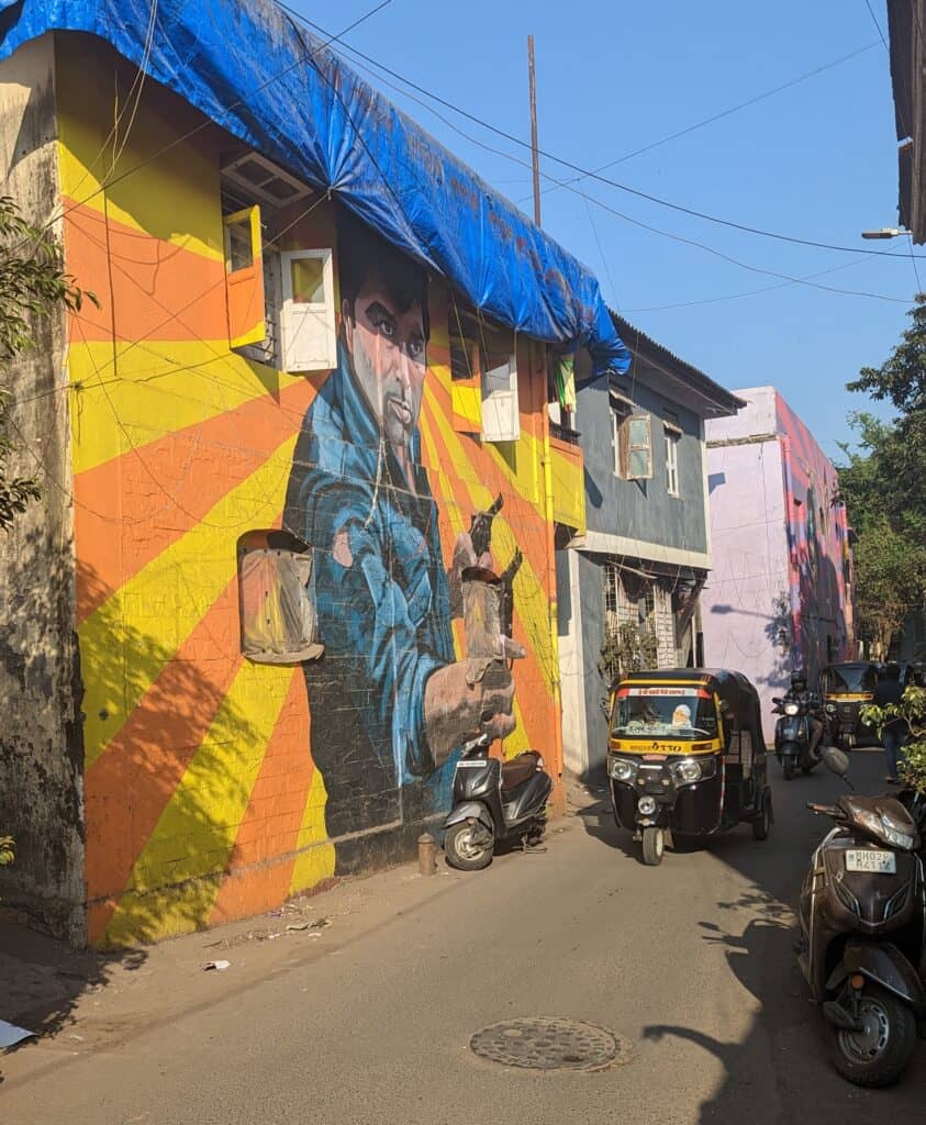 A tiny street in Bandra, Mumbai with a yellow, black auto rickshaw or tuk tuk. There is also a mural of a Bollywood movie from the 70s and many parked two wheelers in the picture.