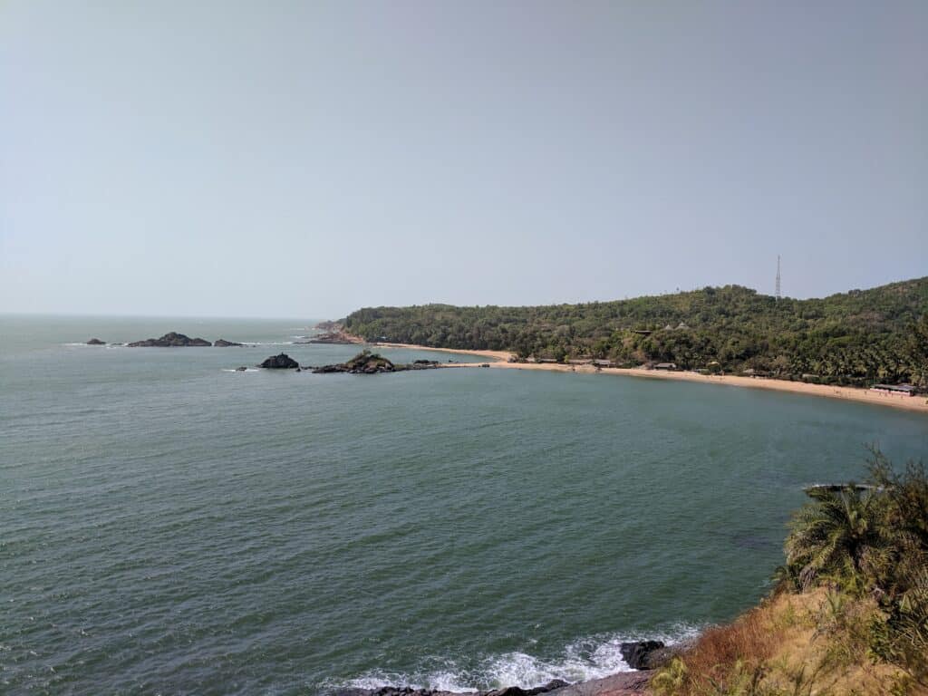 A picture of Om beach in Gokarna India. It is taken from a sea cliff overlooking a long beach with two crescent shapes. There is also a hill that runs along the crescents forming the background. 