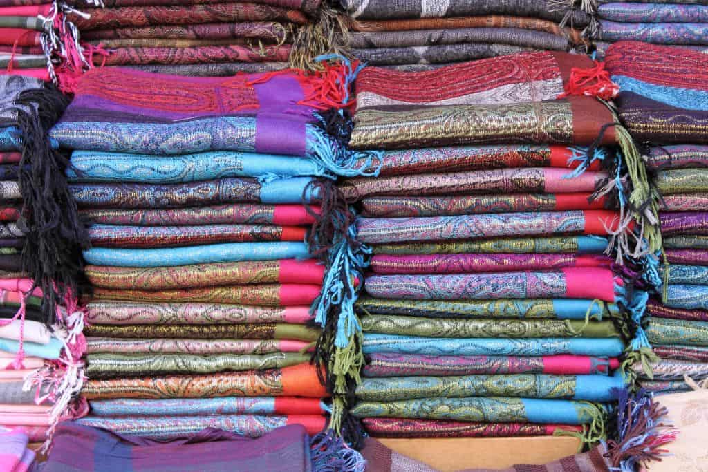 A stack of colorful pashmina shawls neatly arranged in a market stall, showcasing intricate patterns and vibrant hues of blue, red, purple, and green. The soft fabric and detailed designs highlight the fine craftsmanship, with each shawl adorned with delicate fringes, inviting shoppers to explore these luxurious textiles