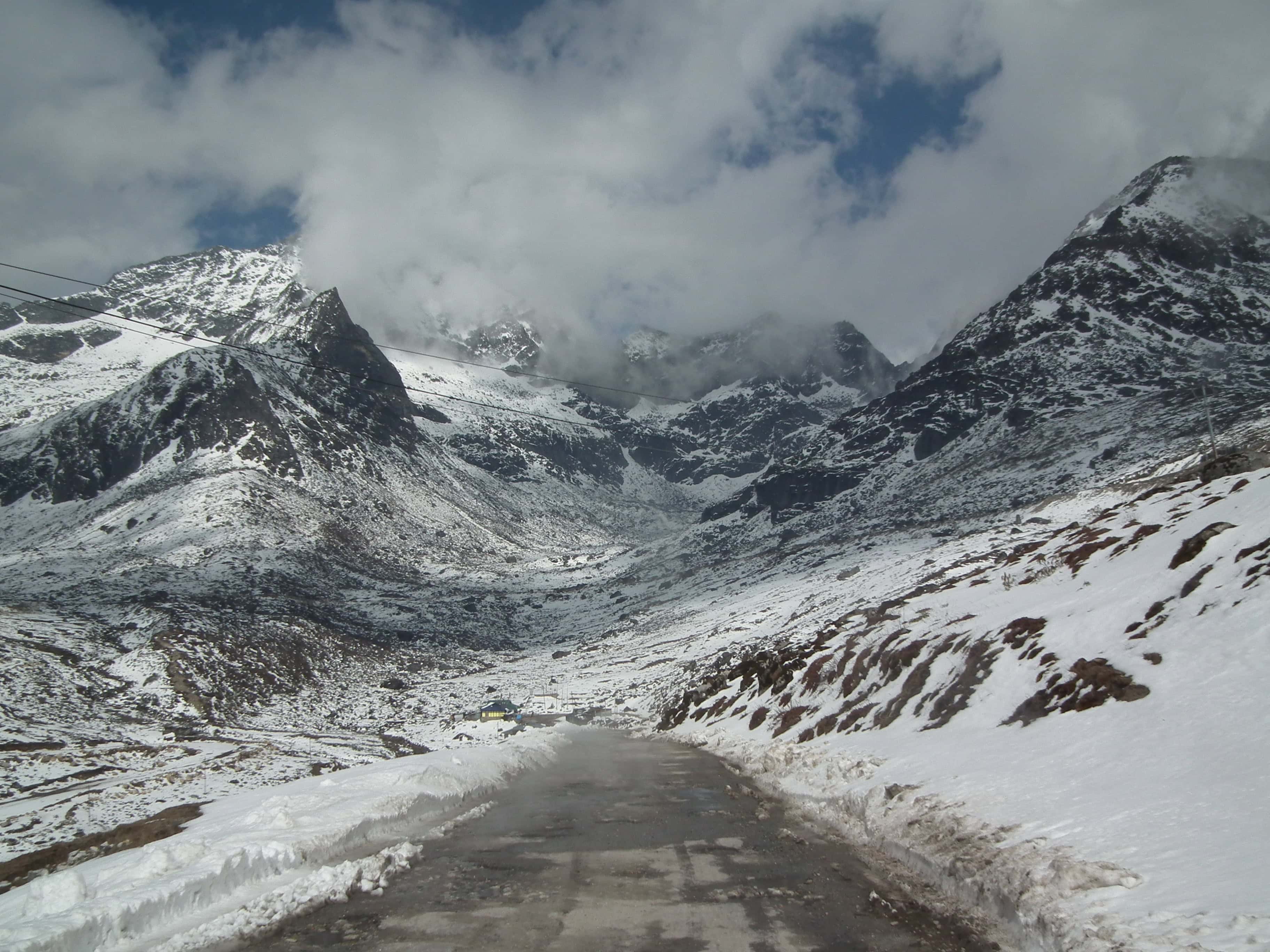 An icy one lane road is in the middle of the picture surrounded by snowy mountain peaks that surround it.