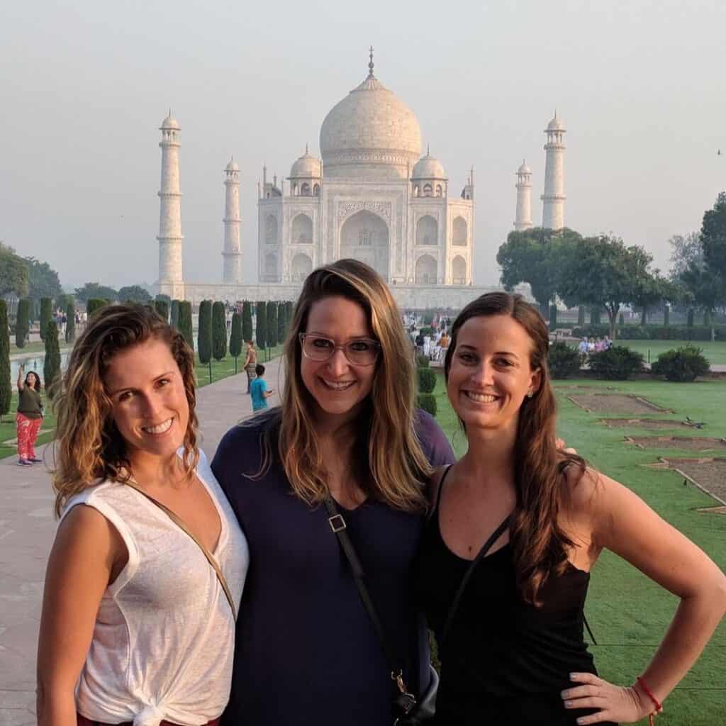 Three smiling women pose for a photo in front of the Taj Mahal in Agra, India. The iconic white marble mausoleum is visible in the background, with its large central dome and minarets. The women, are dressed casually - one in a white top, one in a purple shirt, and one in a black top. They stand on a pathway with manicured gardens visible around them.