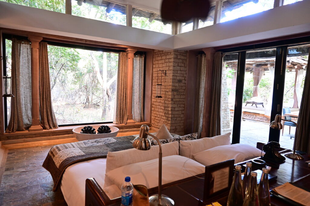 A luxurious stone cabin inside a forest. The picture is taken from inside the cabin and shows a plush bed with glass windows and doors all around the cabin providing forest views. 