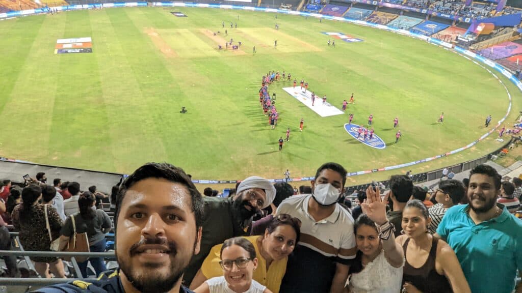 A family at the Wankhede Cricket Stadium in Mumbai. A selfie taken from the stands with cricketers on the field in the background lined up shaking hands after a game. 