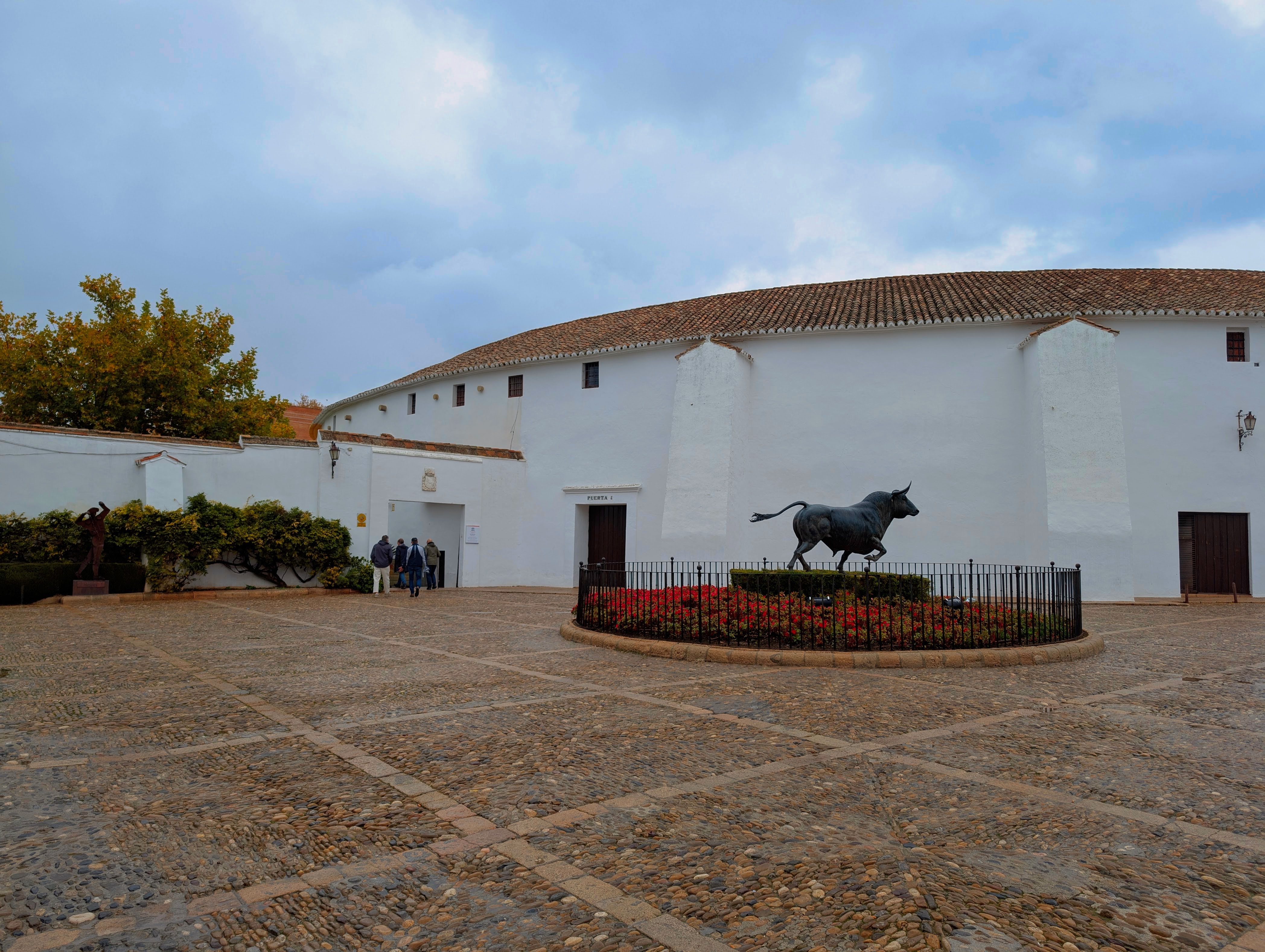 A view of the bull ring in Ronda, Spain. It is the oldest bull ring in Spain, completed in 1785. The picture is taken from outside the arena with the statue of a bull around a flower bed in the foreground. There is an entrance to the bull ring where you see people waiting in a queue. The building itself is circular, with white walls and a red thatched roof. 