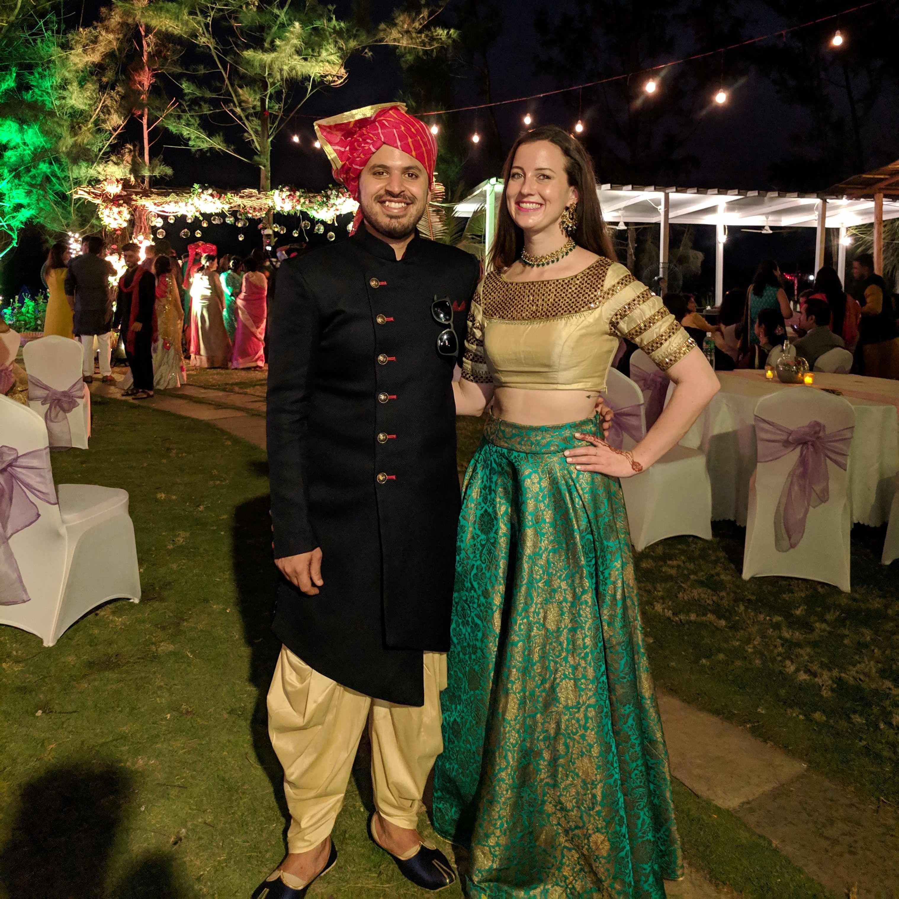 A couple dressed in traditional Indian wedding attire at an outdoor evening celebration. The man wears a black sherwani with beige pants and a red turban, while the woman wears a gold blouse with a teal and gold embroidered lehenga, accessorized with traditional jewelry. The background features decorative lights and wedding guests.