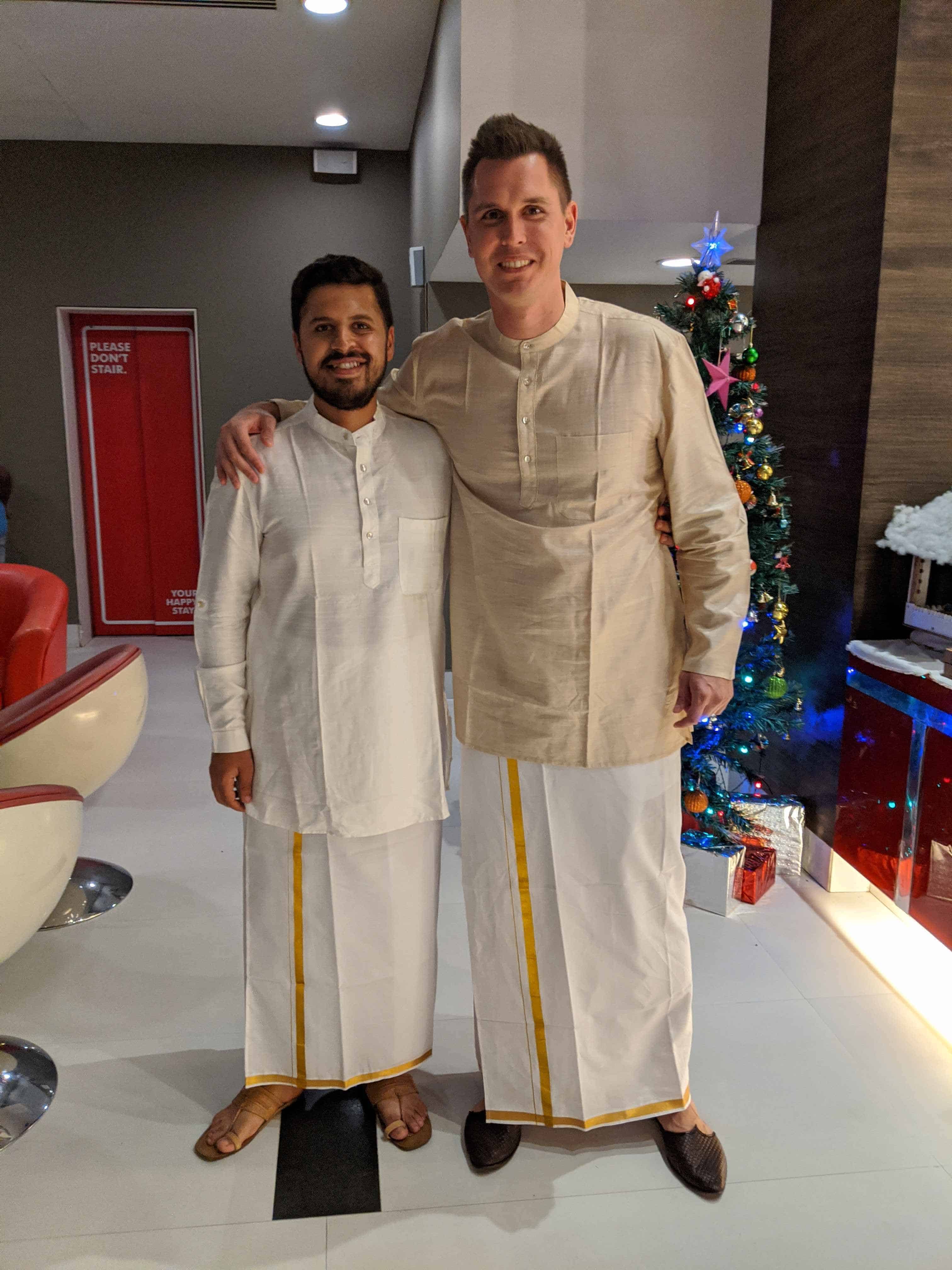 Two men standing together and smiling, dressed in traditional Indian attire consisting of off-white shirts and dhotis with gold borders. They are indoors near a Christmas tree decorated with ornaments, with festive decor in the background.