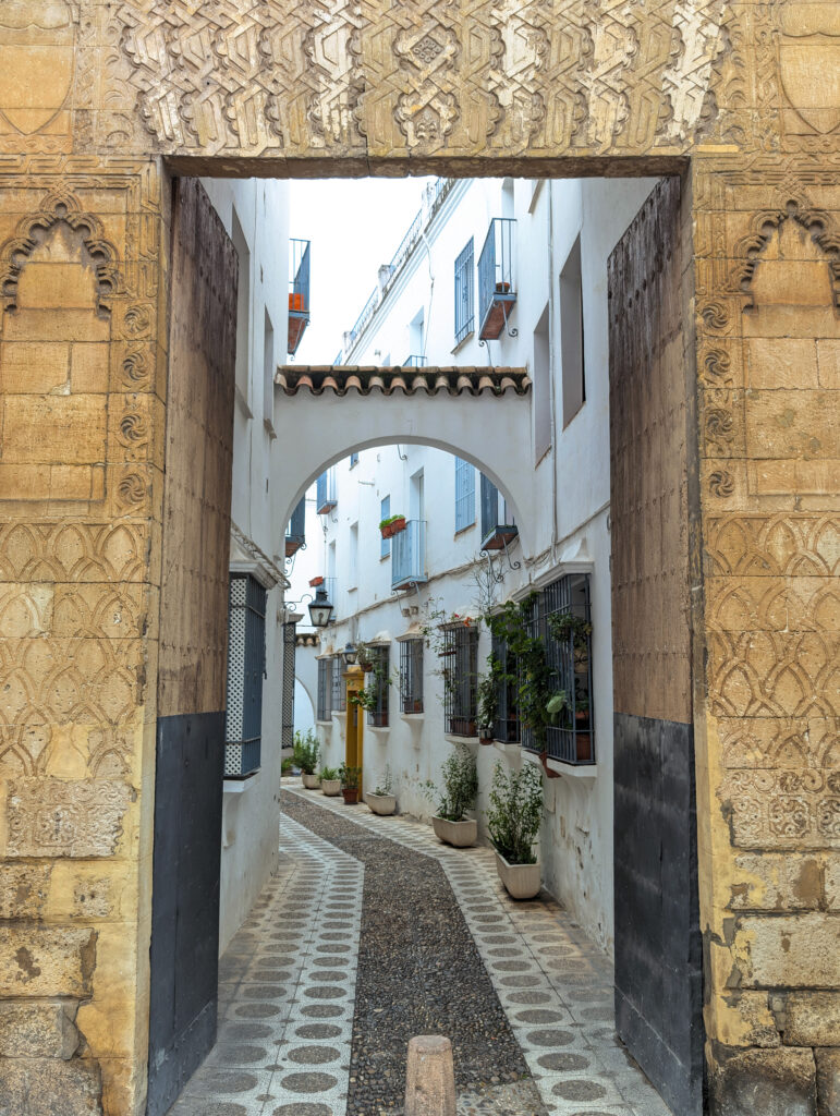 A picture of an alley in an old town in Cordoba, Spain. There is an Islamic door with an arch in the background as the alley narrows. There are buildings on either side of the alley painted in white with blue windows. 