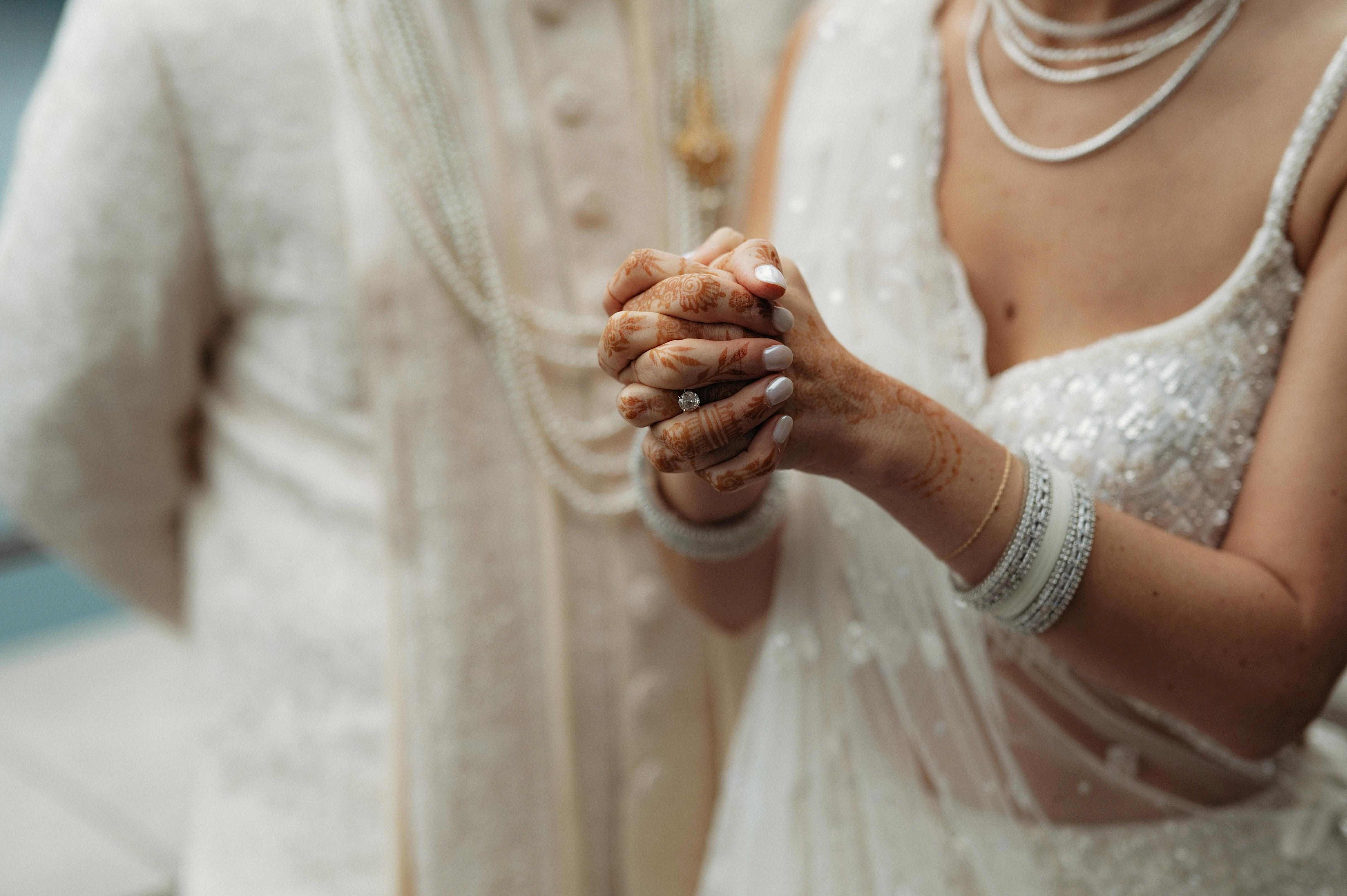 A close-up of a couple's wedding outfit details. The bride's hands are folded in front and feature intricate henna designs and a solitaire engagement ring, while her nails are painted a soft, neutral color. She is wearing an elegant white lehenga with shimmering details and layered pearl necklaces, along with matching white and silver bangles. The groom, dressed in a light-colored traditional outfit, is partially visible in the background.