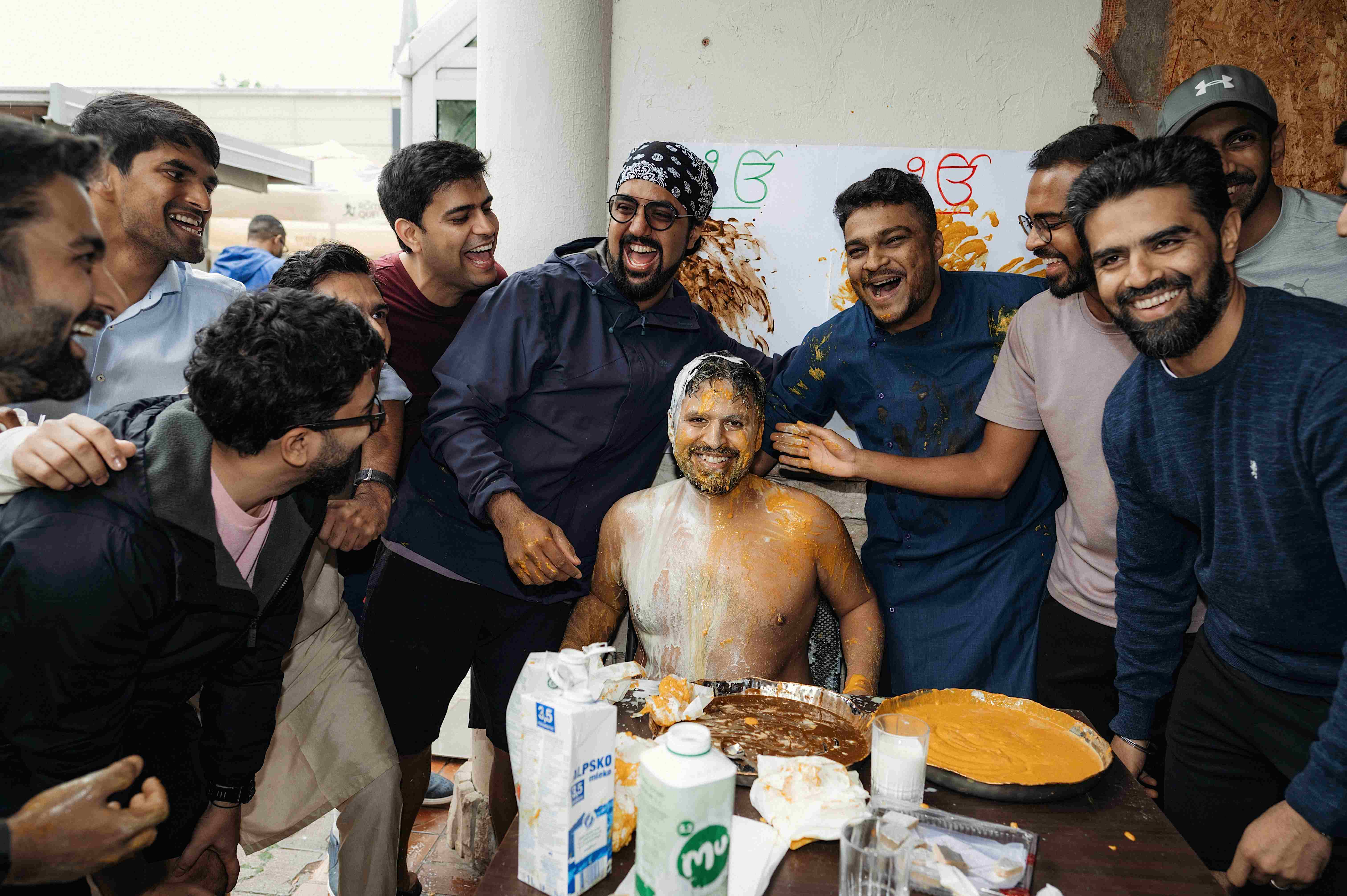 A group of men smiling and laughing during a lively Indian wedding Haldi ceremony. The groom, sitting shirtless and covered in turmeric paste, is surrounded by friends applying more turmeric in a playful and joyous atmosphere.