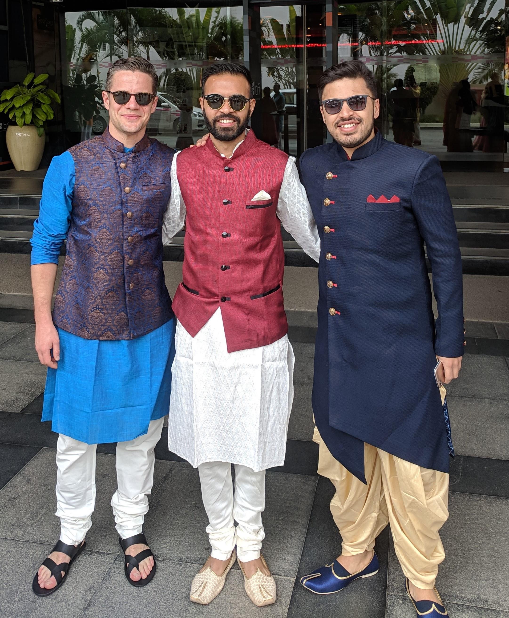 Three men dressed in traditional Indian wedding attire, smiling and posing together. Each wears a distinct outfit: one in a blue kurta with a brocade waistcoat, another in a red and white kurta set with mojari shoes, and the third in a navy sherwani with gold dhoti pants and matching juttis, all accessorized with sunglasses.