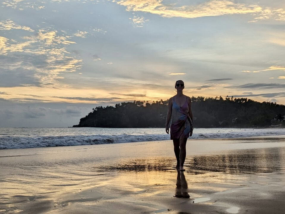 A silhouette of a woman walking along a beach during a colorful sunset at Mirissa Beach. The sky is filled with beautiful golden and purple hues, casting a warm glow over the tranquil ocean. In the background, you can see a hill outcropping with palm trees, further adding to the serene and picturesque setting. The person's reflection can be seen clearly in the wet sand, creating a mirror-like effect.