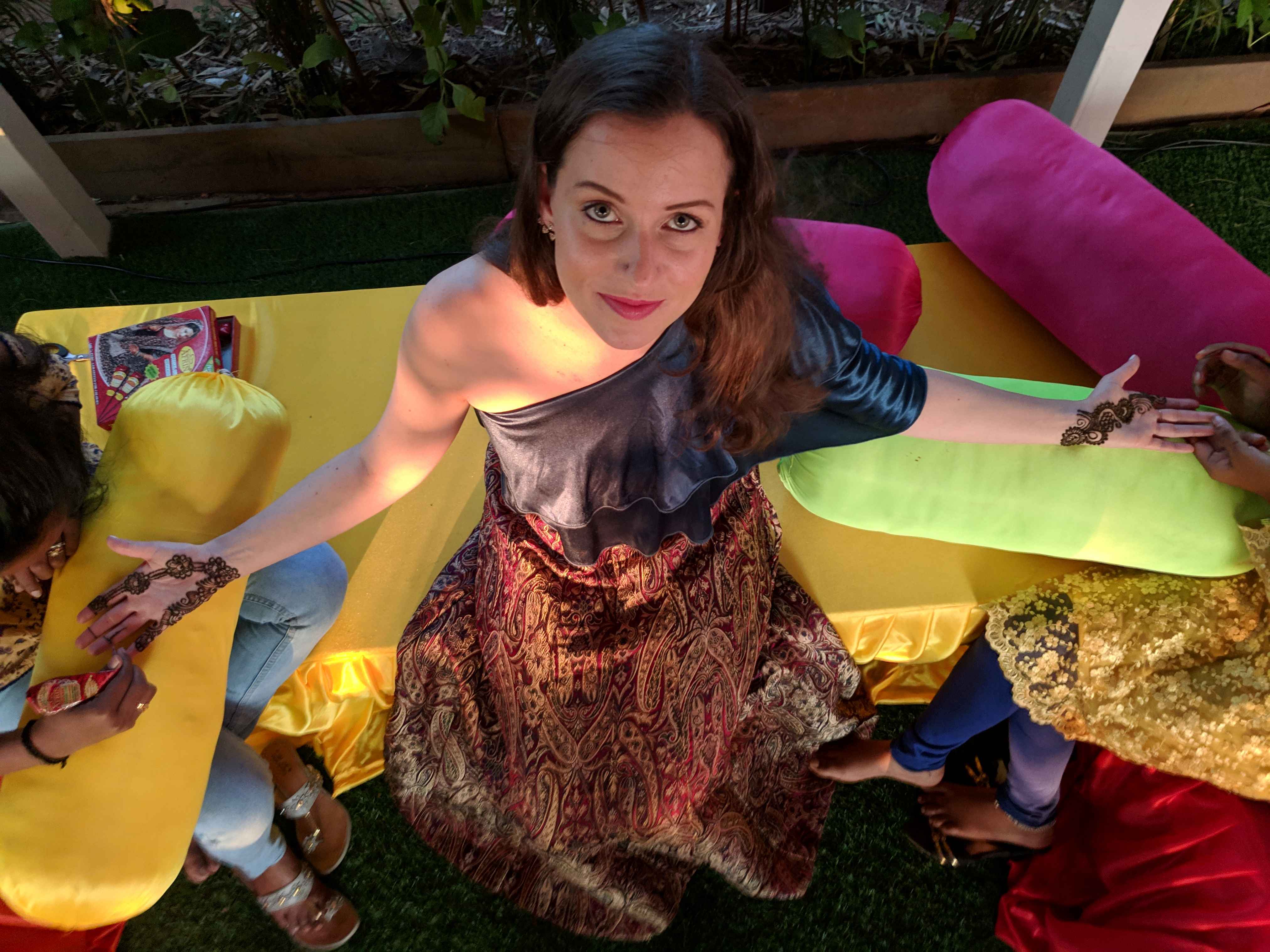 A woman sitting on a colorful cushion setup at a mehndi ceremony, having intricate henna designs applied to her hands. She is dressed in a one-shoulder blue top paired with a patterned skirt, surrounded by bright yellow, green, and pink cushions, creating a festive atmosphere.