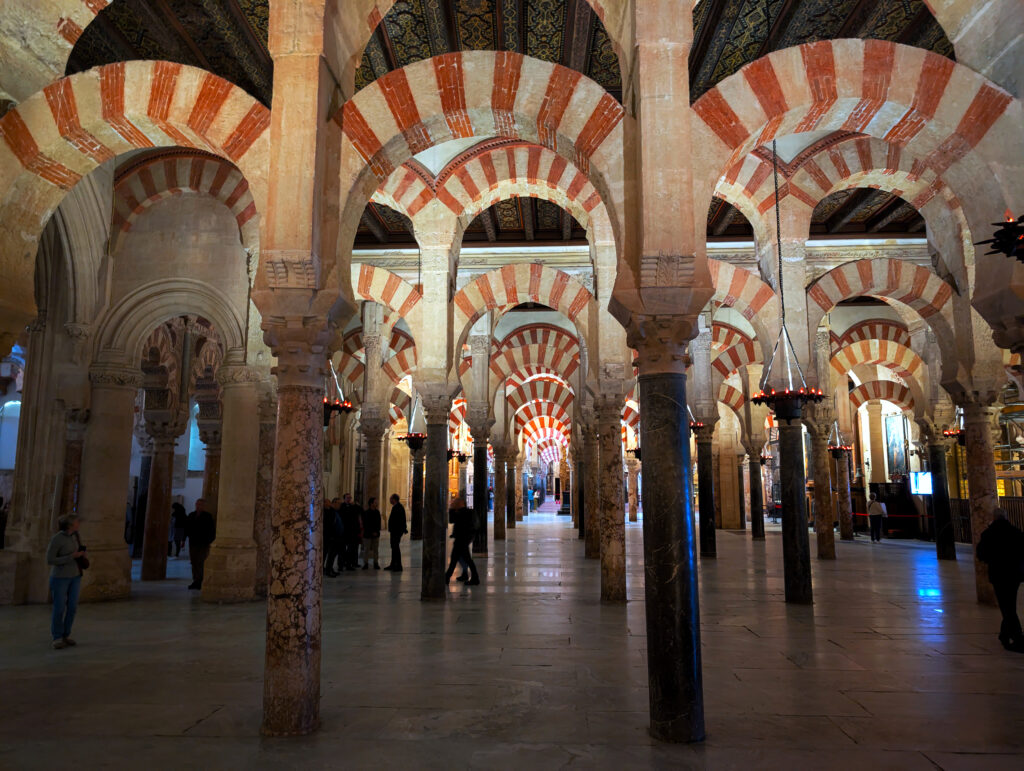 Endless arches in a dimly lit hall at the Mosque-Cathedral monument in Cordoba, Spain. The building was built as a mosque over 1000 years ago and was converted into a cathedral. The picture is of the grand hall with arches painted in white and red stripes that run all along the hallway along ornate marble pillars. There are people walking around admiring the structure.