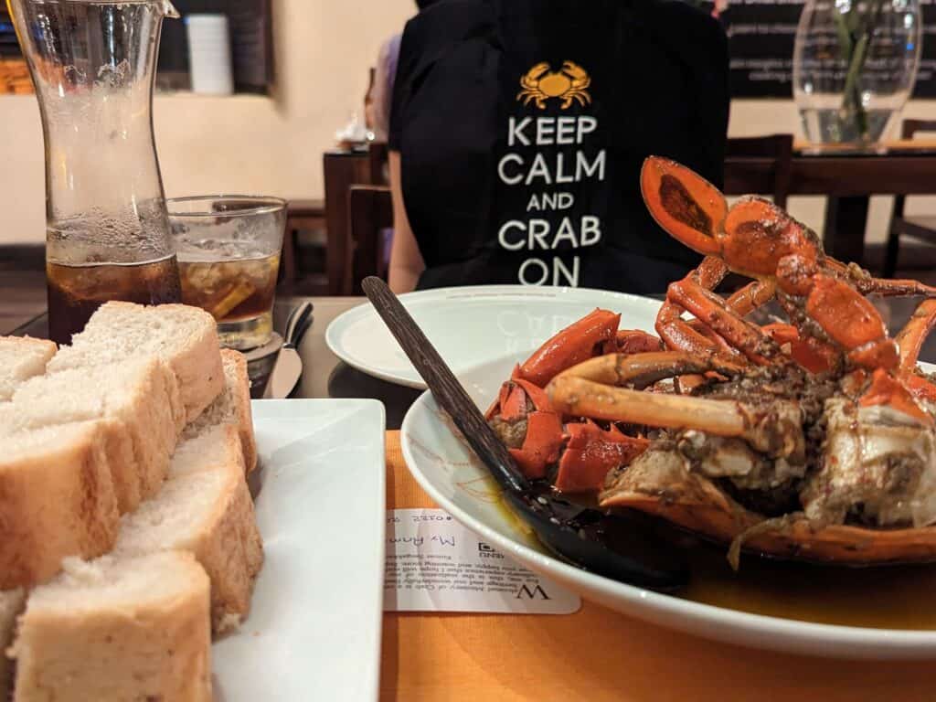 A plate of succulent crab served in a flavorful sauce at Ministry of Crab, Colombo, Sri Lanka, accompanied by slices of bread and a glass of iced drink. A diner in the background wears a bib with the words 'Keep Calm and Crab On.'