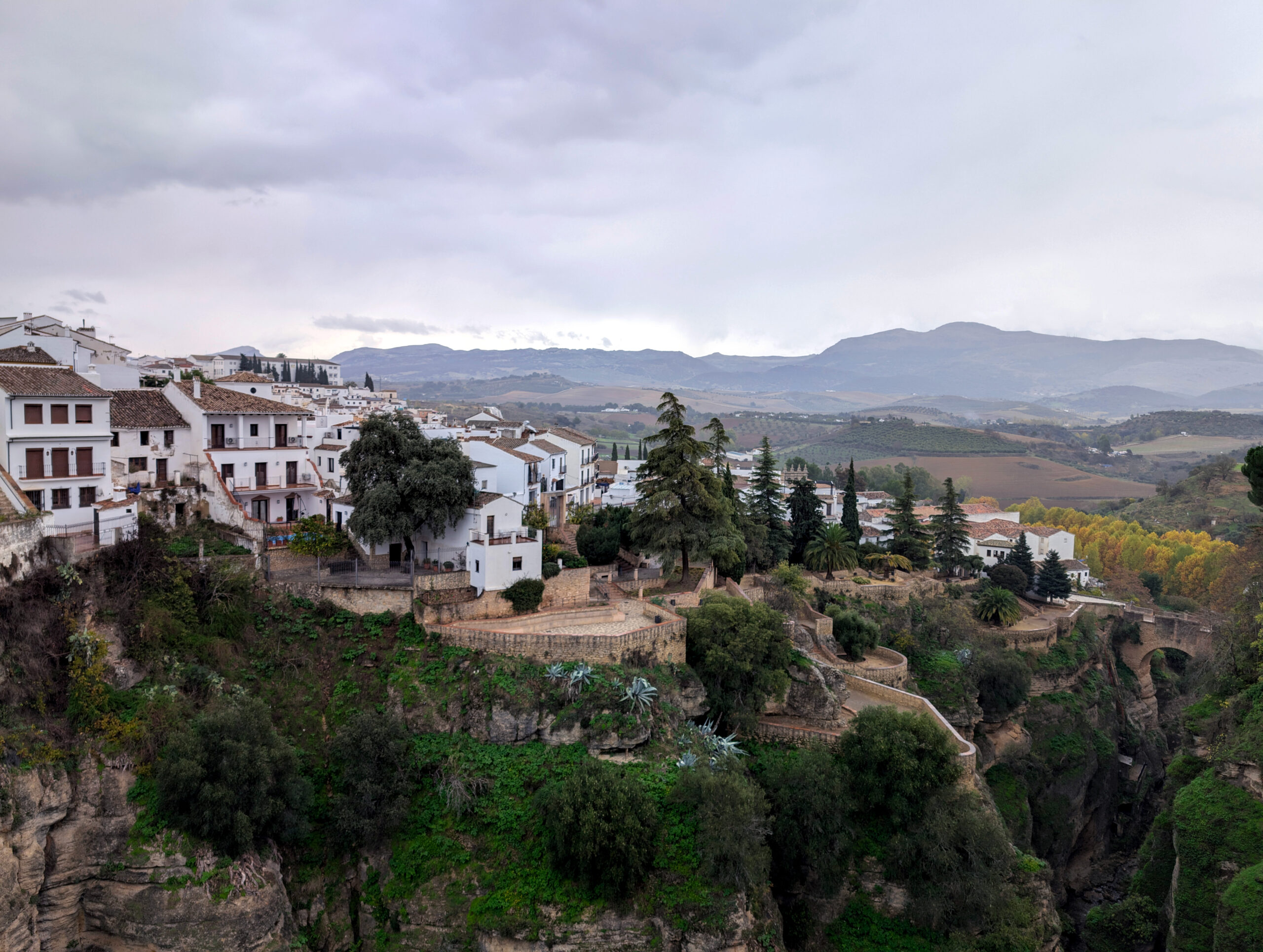 A view of the old town in Ronda, Spain. It is set over a deep gorge with vertical rock cliffs. The picture shows the old town with white buildings, red roofs and medieval walls leading a path down to a 16 century stone bridge. 