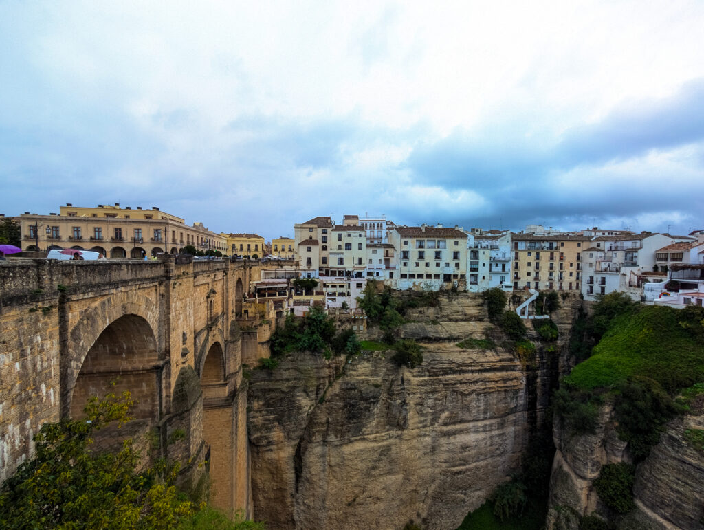 A picture of the Puente Nuevo bridge in Ronda, Spain. The picture shows two arches of a bridge built on a canyon with town buildings painted in white, yellow and blue in the background. The picture has been taken on a rainy day and there are storm clouds in the sky. 