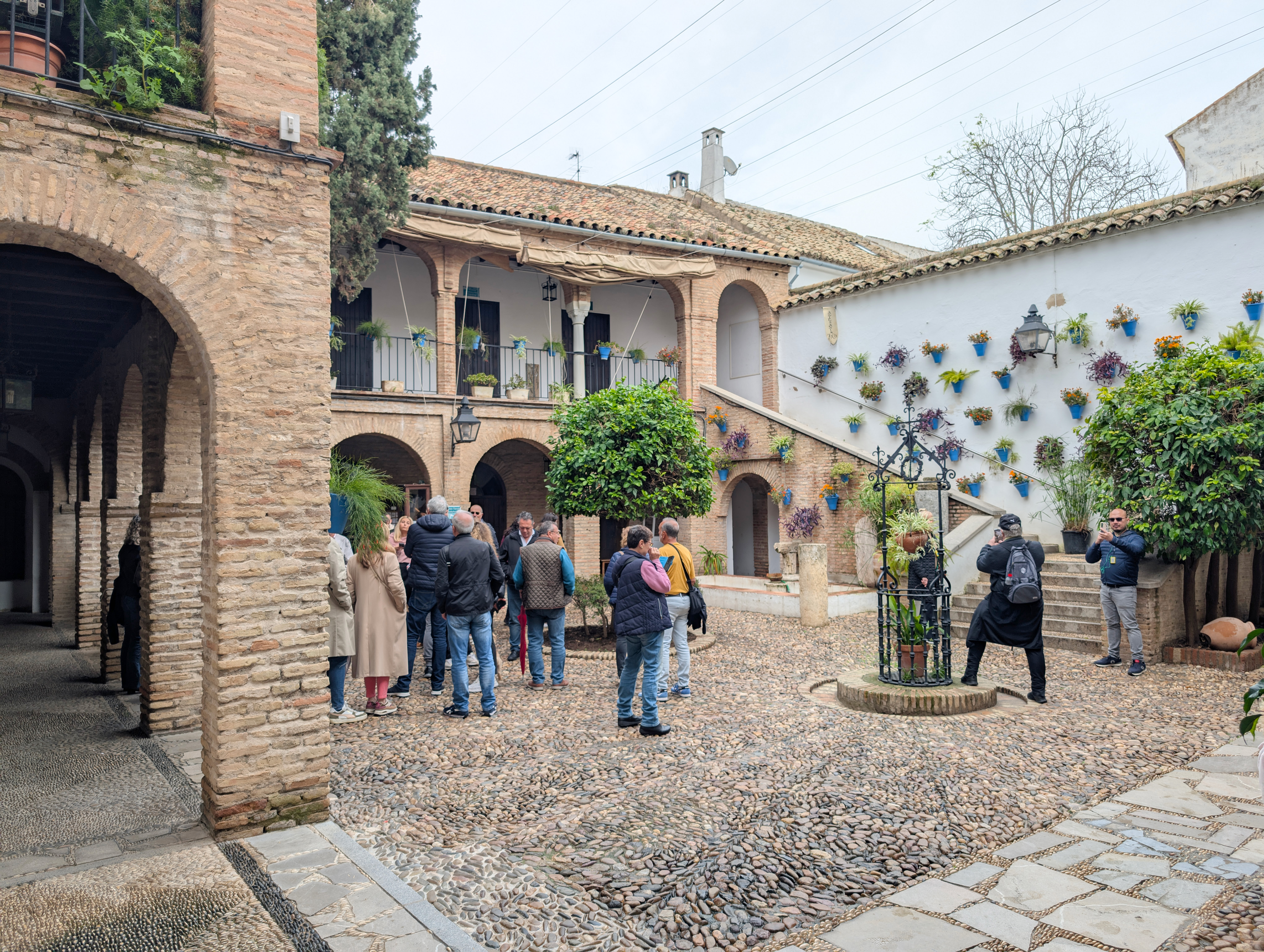 A picture of a traditional Cordoban patio in Spain. THe patio has blue flower pots all along the walls and arched corridors. There are also orange trees in the middle of the patio with tourists all around the patio taking pictures. 