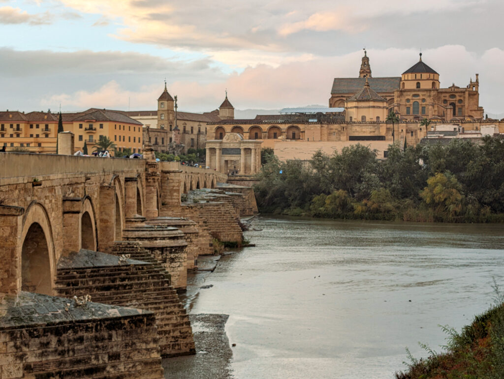 A view of the Roman Bridge in Cordoba, Spain. The bridge is a medieval bridge first built in the 1st Century by the Romans. It is made of brownstone with arches. There is a church and a medieval old town in the background. The sky can be seen at sunset with clouds scattered with orange, red and blue hues.