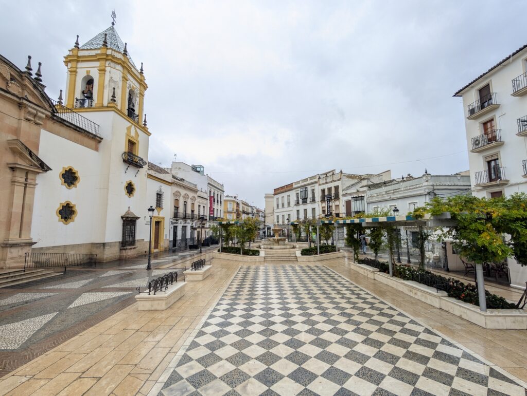 A view of the main square in Ronda's old town in Spain. Plaza del Socorro has a yellow and white church on its left with many restaurants and buildings along the square. The buildings are white and the floor is checkered. It is a rainy day and there are people with umbrellas walking around. There are also little orange trees along the right edge of the square, something common across the Spanish province of Andalusia. 