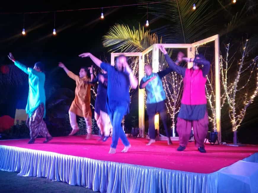 A group of men in colorful traditional Indian attire dancing energetically on a small decorated stage at night, with string lights and a festive backdrop of illuminated trees and palm leaves.