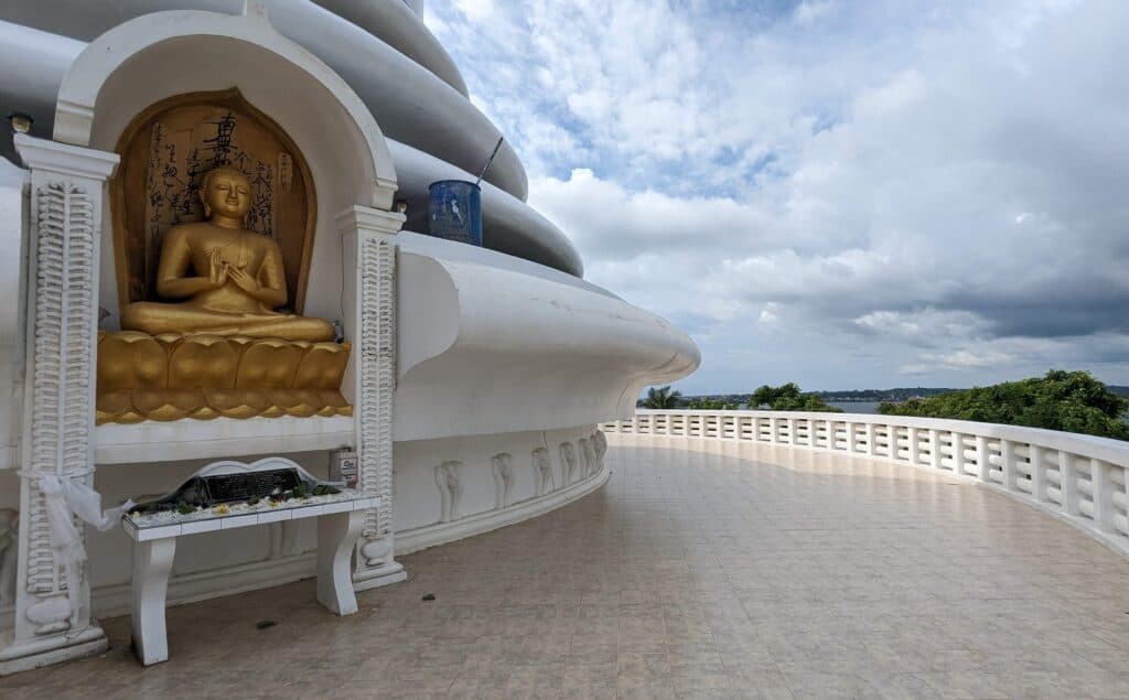 The exterior of a large white Buddhist stupa or pagoda structure located in Unawatuna, Sri Lanka. The centerpiece of the structure is a large gilded Buddha statue seated in a meditative pose within an arched alcove. The pagoda is surrounded by a wide, tiled balcony with ornate white railings that offer expansive views of the lush, green surroundings. The sky overhead is filled with dramatic, cloudy formations.