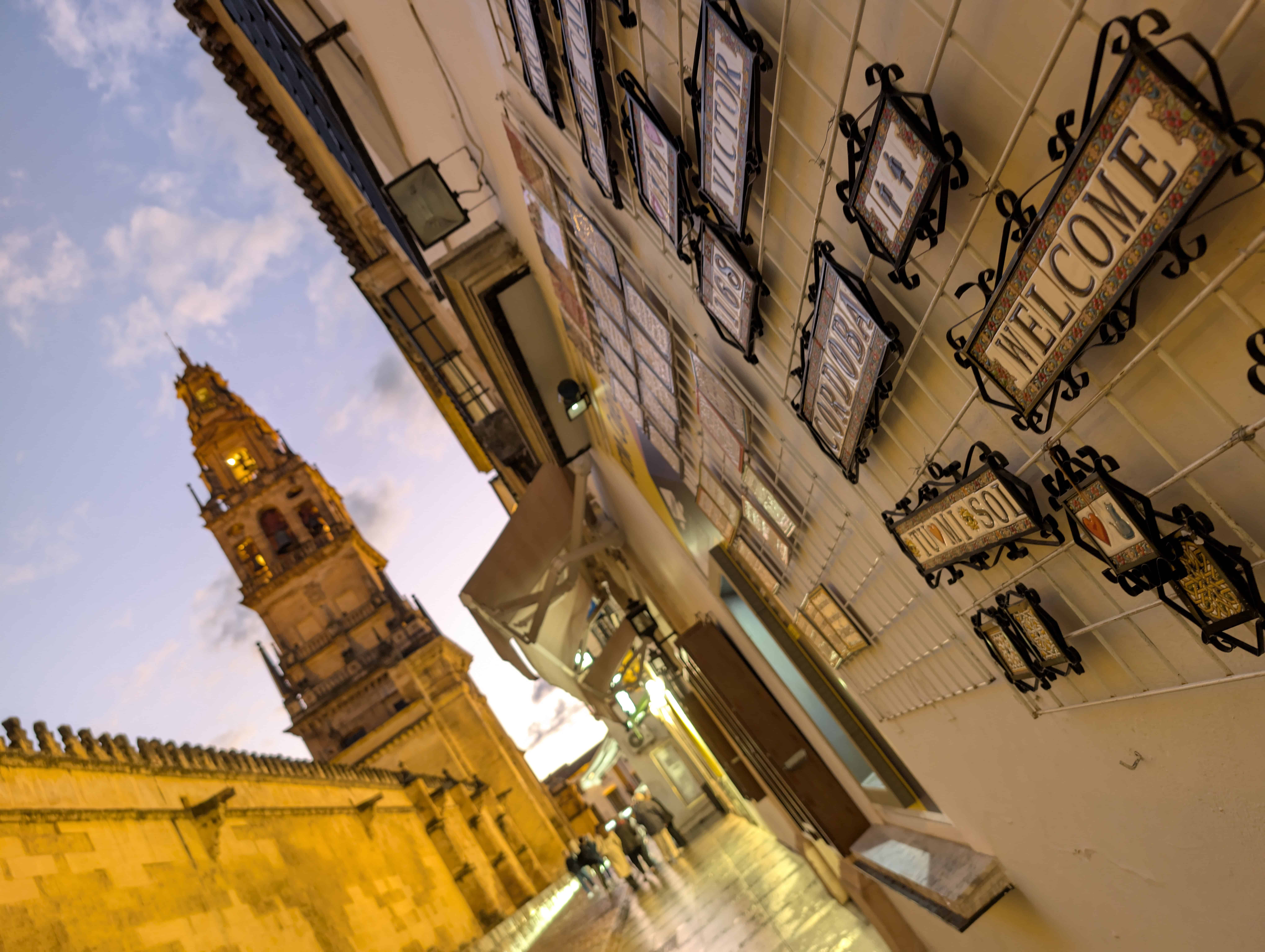 A view of a medieval bell tower in Cordoba, Spain. The picture has been taken at dusk and the sky in the background is radiant, with the walls and tower lit up in yellow light. You can also see souvenirs along the walls of shops on the street. 