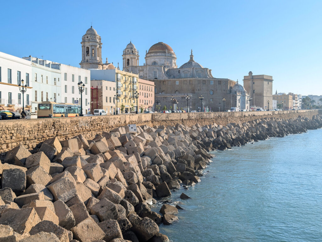 A view of the Cadiz Cathedral in the Spanish city of Cadiz in Andalusia. The picture has been taken from near the sea and you can see a sea wall with rocks and a promenade, road that runs along it. The imposing cathedral can be seen in the background with other colorful buildings. The dome of the cathedral is red with white stone around it. The building also has two distinct bell towers. 