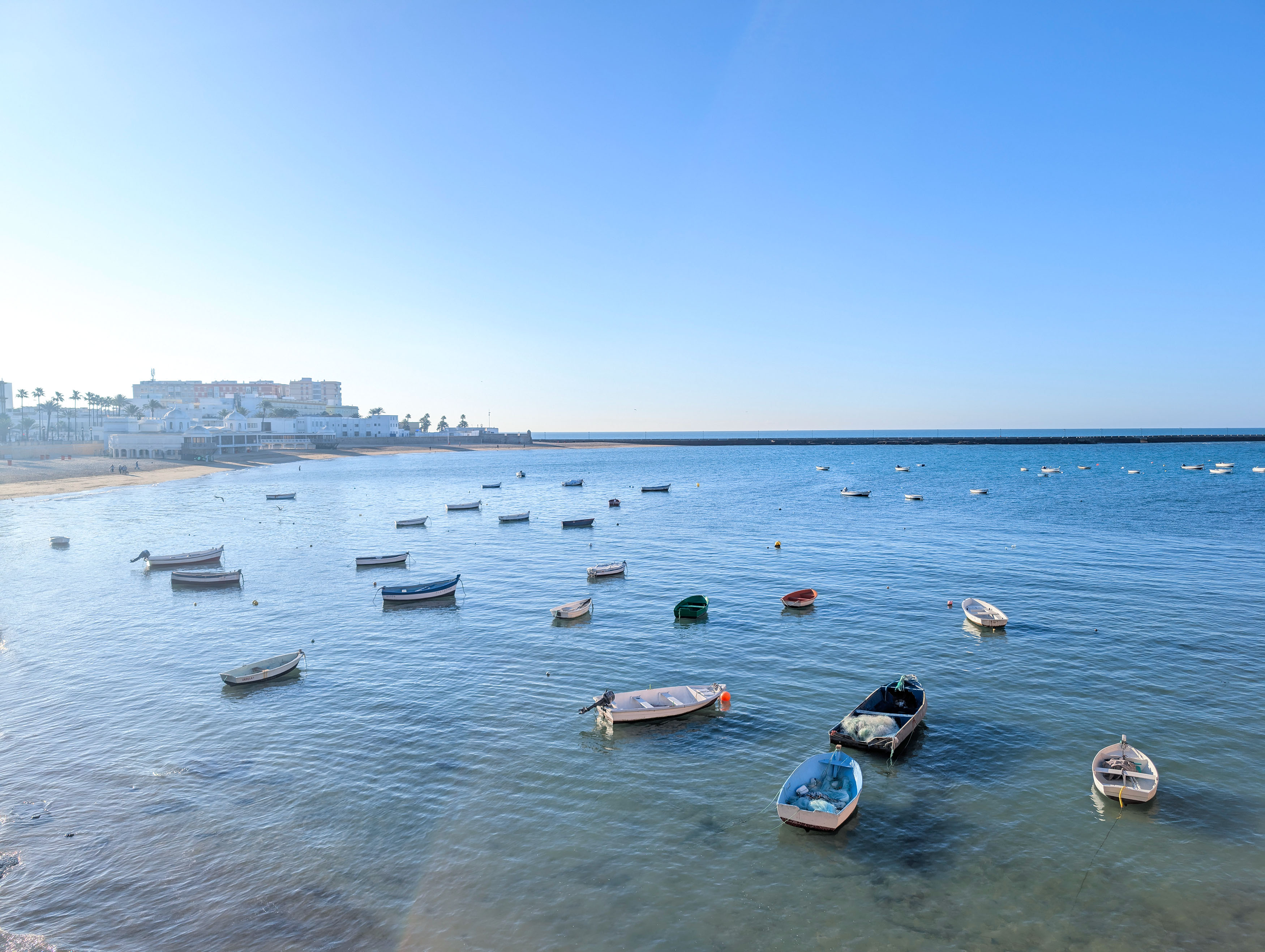 A view of the sea and beach in Cadiz in Spain. There are many boats on the blue water. The picture has been taken at sunrise and you can see the beach with the city in the backdrop. 