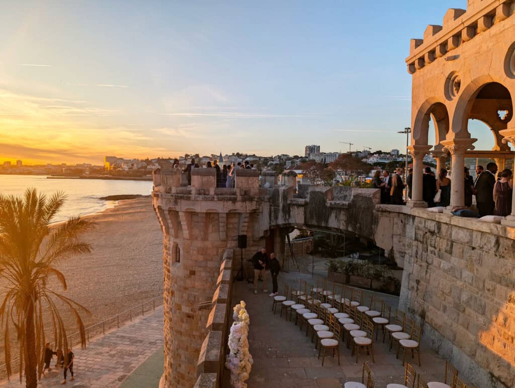 Scenic view of a historic castle terrace in Estoril, Portugal, at sunset, with people enjoying the golden hues over the Atlantic Ocean. The beach, palm trees, and elegant seating add to the romantic and serene atmosphere of the coastal town.