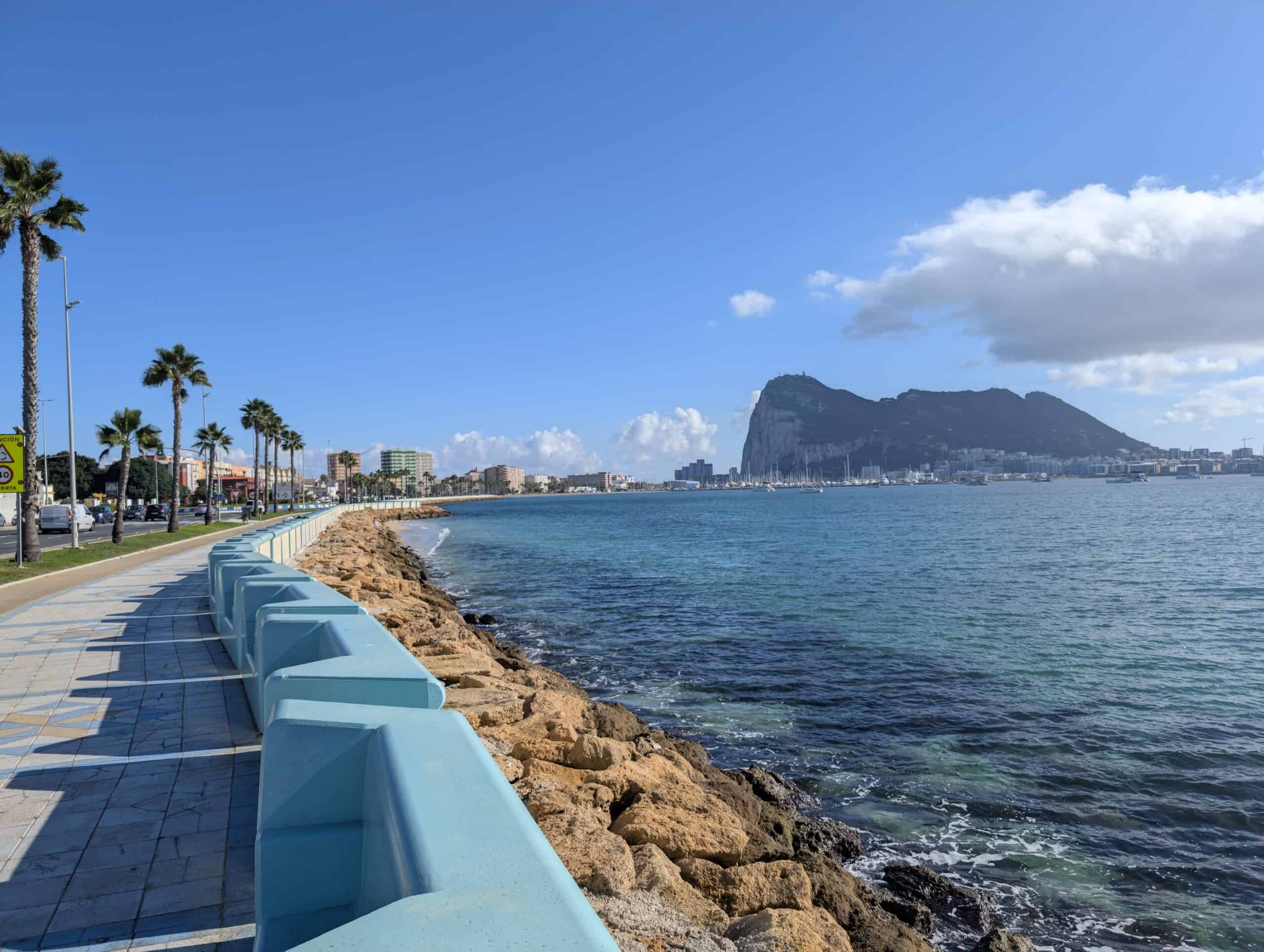 A view of the Rock of Gibraltar and the blue waters of the strait. The picture has been taken on a seaside promenade with palm trees lining its left. The picture also shows the city of Algeciras in Spain. 