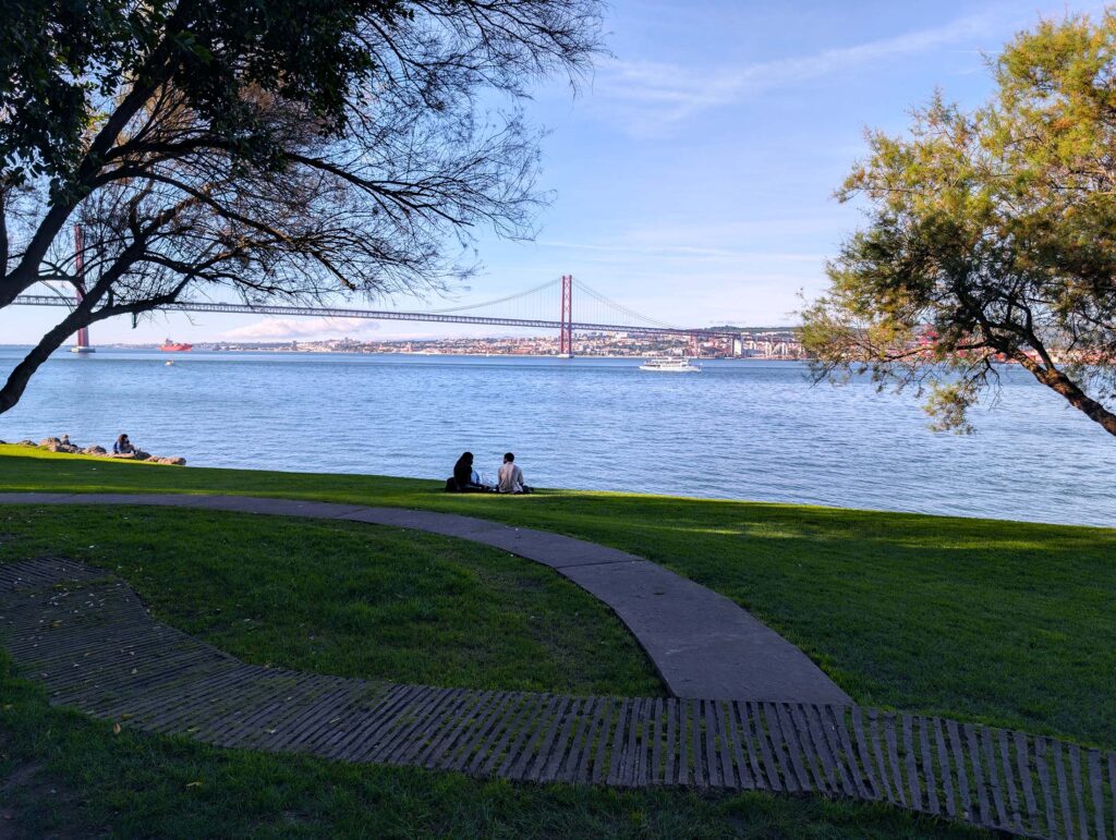 Scenic view from Jardim Cacilhas in Almada, Portugal, showcasing a green park with shaded trees and people relaxing by the Tagus River. In the background, the iconic 25 de Abril Bridge spans across the water, framed by a clear blue sky.