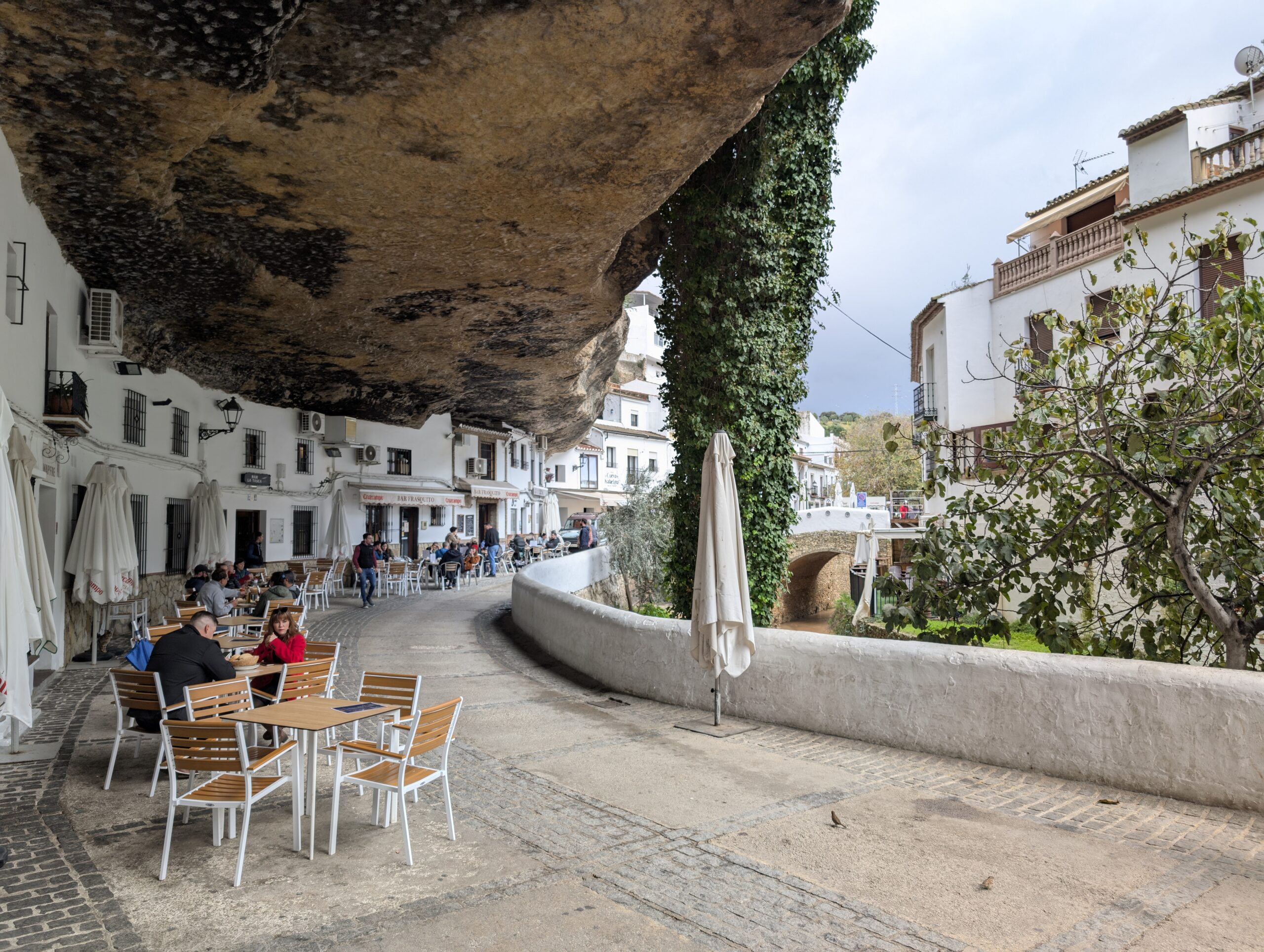 A picture of a street in Setenil de Las Bodegas in Andalusia, Spain. The street is cut into a rock face by a stream. There are little white buildings that serve as shops and restaurants along the left edge of the picture. You can also see tables with tourists eating lunch under the carved rock face. There is a rock ceiling to the street that can be seen along the top of the picture. 