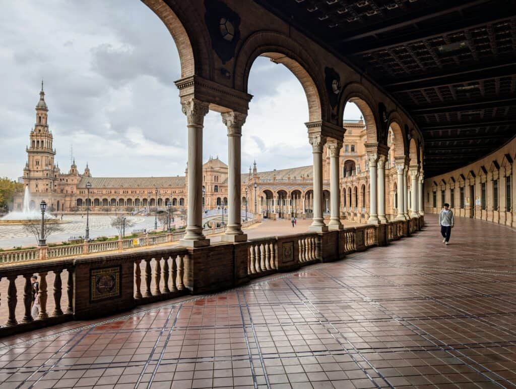 A picture of the stunning Plaza de Espana in Seville, Andalusia, Spain. The picture has been taken from a raised walkway by the crescent shaped square in Seville.  The periphery of the crescent has a crescent shaped red palatial building with 3 bell towers. The picture shows almost half the building with ornate arches that run along its entire edge. There is also a water channel and a fountain in the plaza seen in the picture. 