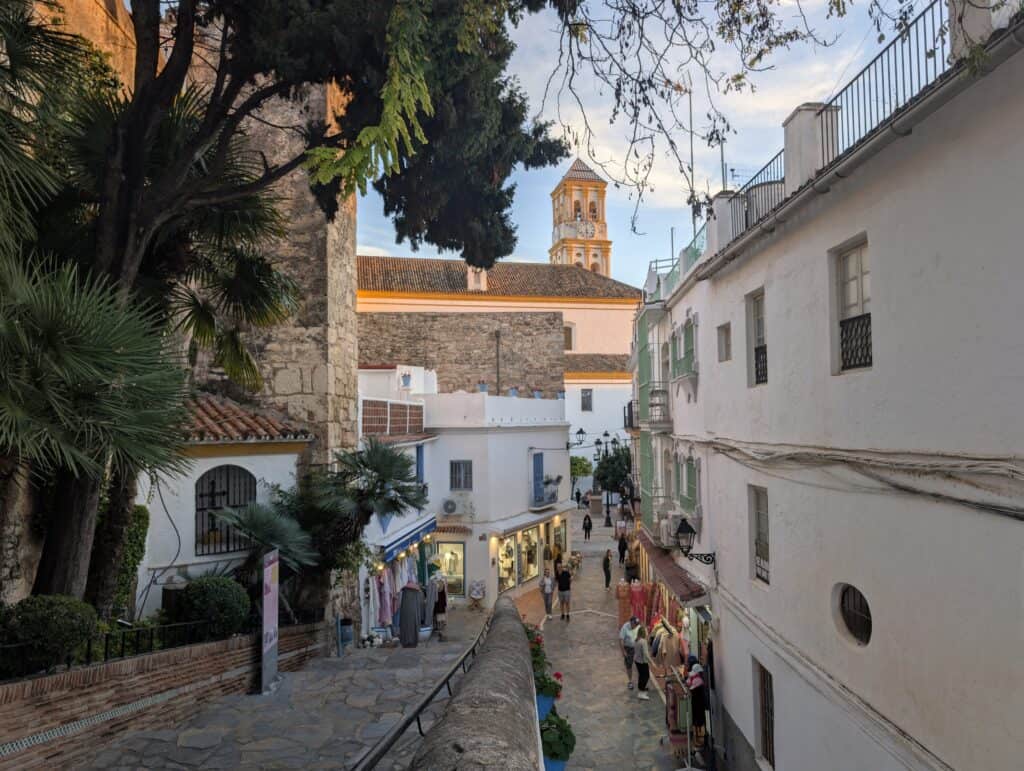 A picture of Marbella's old town in Andalusia, Spain. The picture has been taken in the evening with sunset hues. You can see a staircase and an alley with a church tower in the background. The church is white and yellow in color while the streets of the old town have cobble stones and white walls.