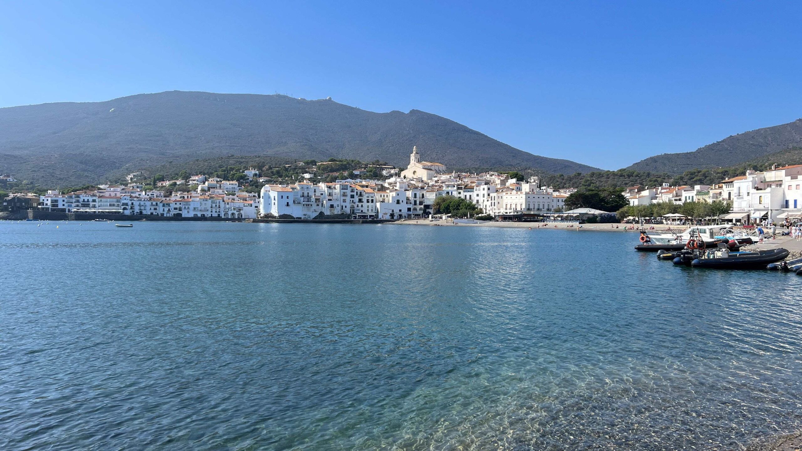 A picturesque coastal view of Cadaqués, Spain, with clear blue waters in the foreground and whitewashed buildings with terracotta roofs lining the shore. Small boats are docked along the beach, and people can be seen enjoying the waterfront. The Church of Santa Maria stands atop a hill, overlooking the town, with green hills and rugged mountains forming a scenic backdrop under a bright blue sky.