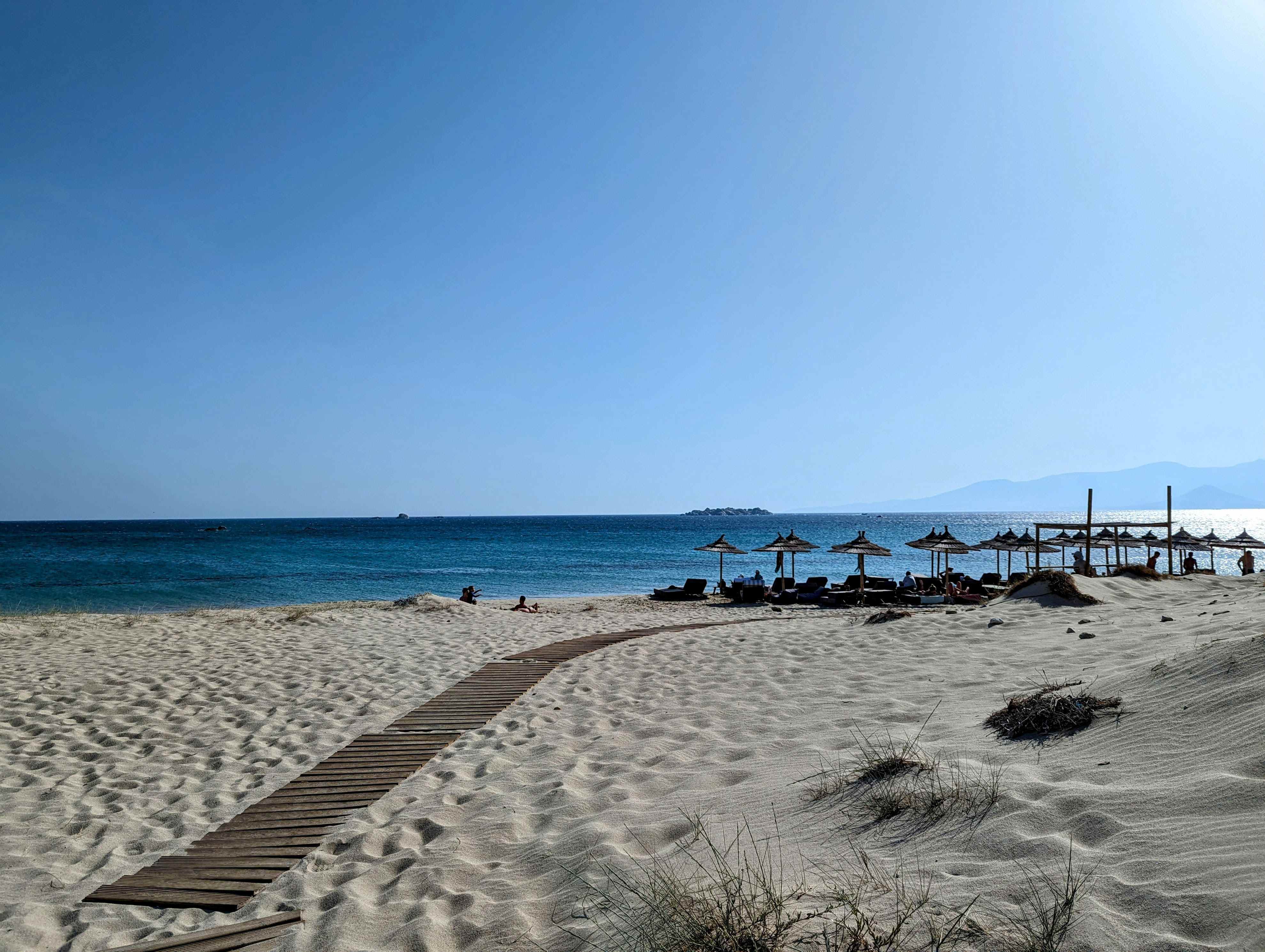 Sandy beach in Naxos, Greece with wooden boardwalk leading to rows of thatched umbrellas and loungers along turquoise waters. Mountains visible in the distance under clear blue skies.