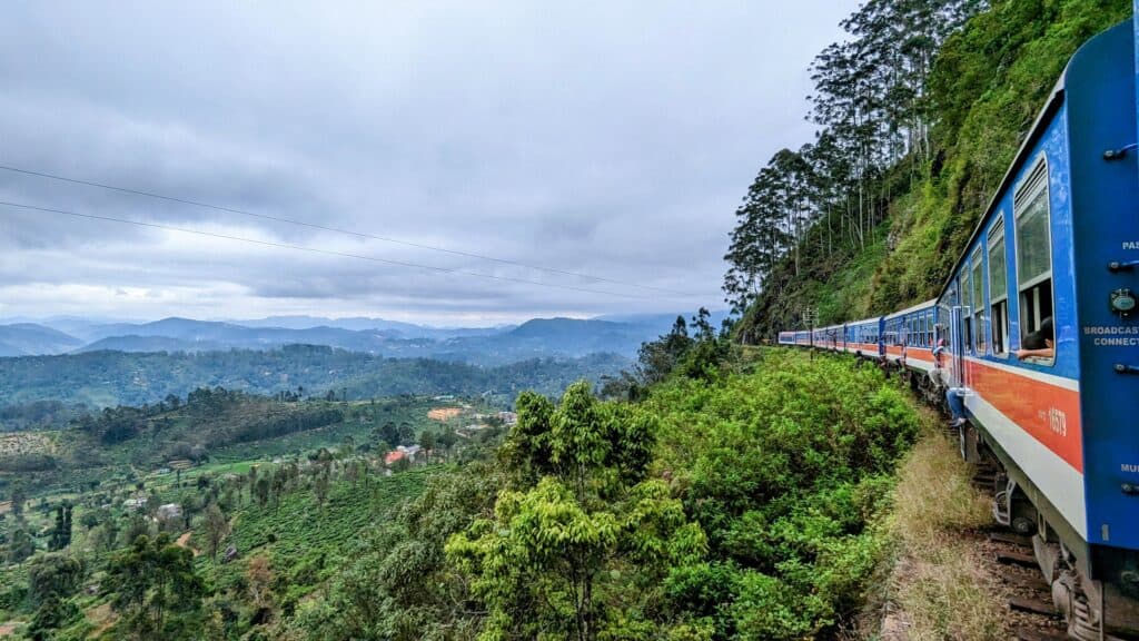 A scenic blue train travels through the lush green hills of Sri Lanka, winding along the mountainside. The train's windows are open, with passengers leaning out to enjoy the breathtaking view of rolling tea plantations, misty mountains, and dense forests under a cloudy sky.